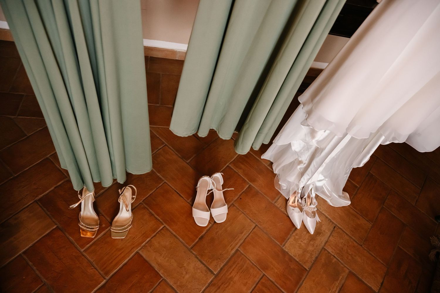 Three pairs of elegant wedding heels displayed on terracotta tile floor beside sage green curtains and a white bridal gown.
