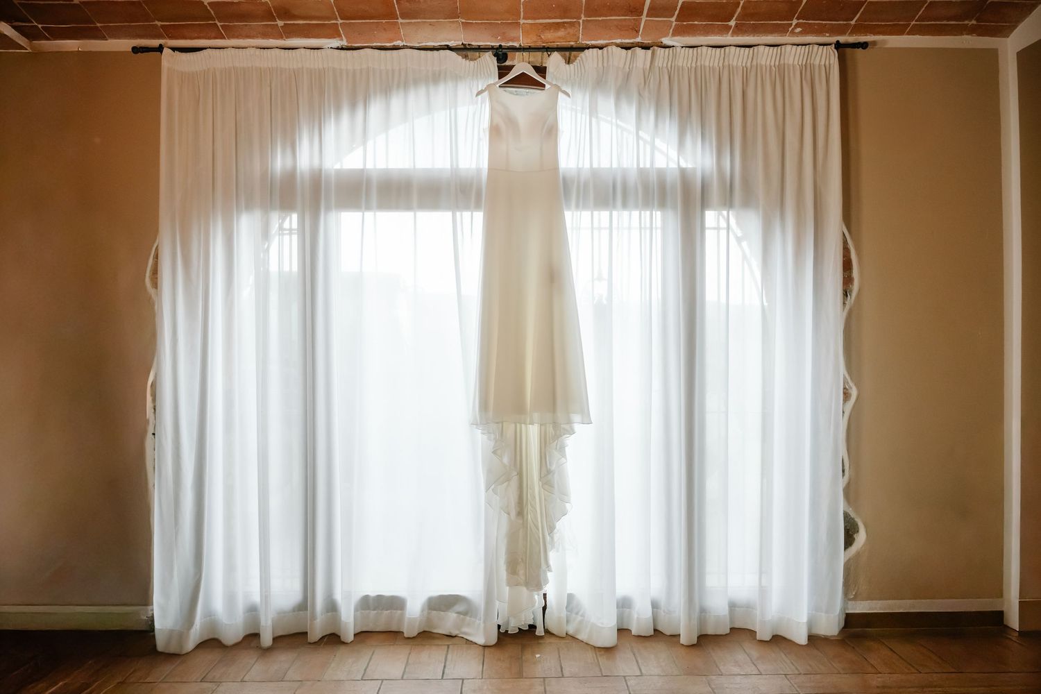 A elegant white wedding dress hanging on a hanger in front of sheer white curtains in a warmly lit room.