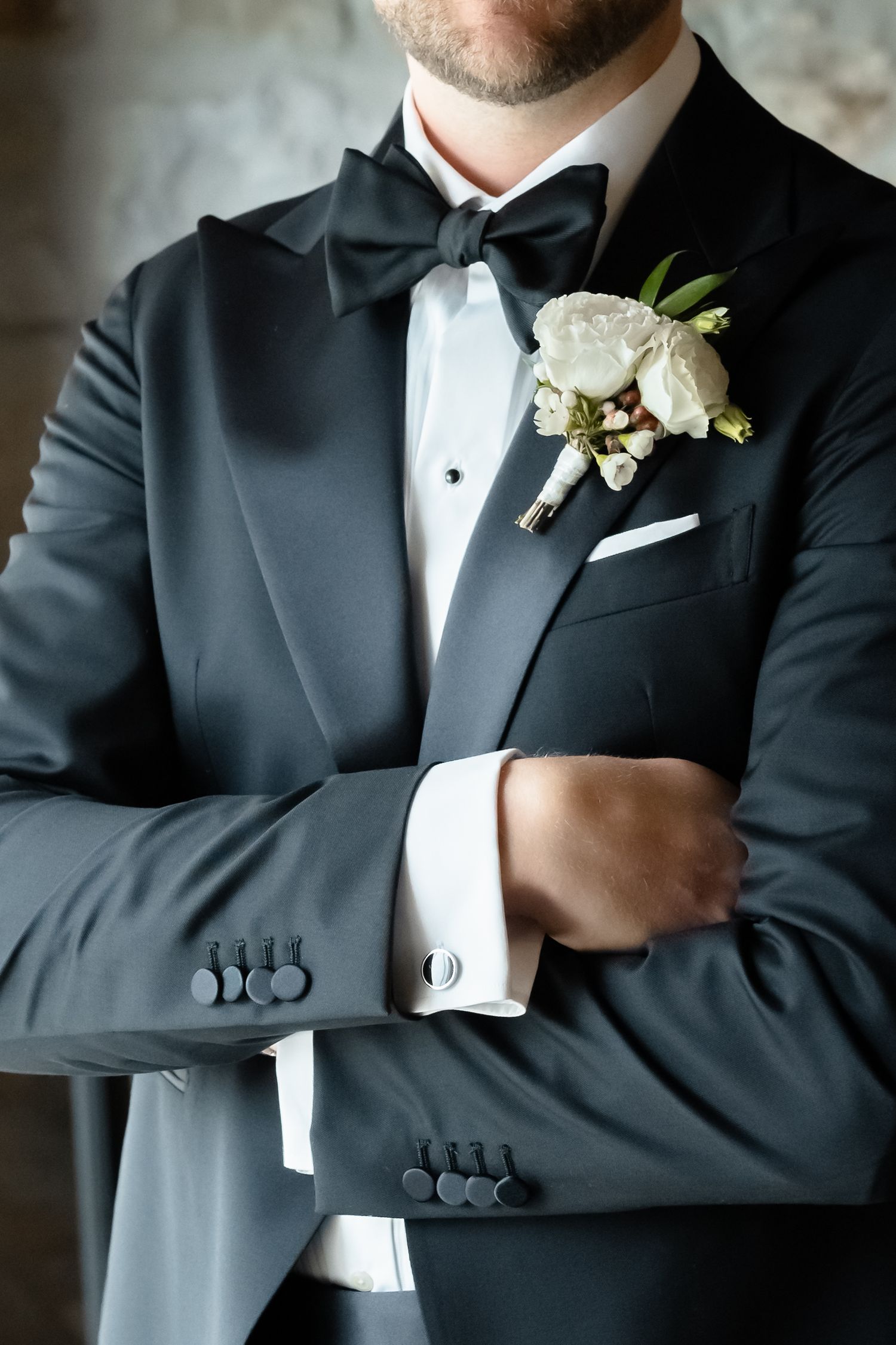 Groom in black tuxedo with bow tie, white boutonniere, cufflinks, and pocket square, arms crossed.