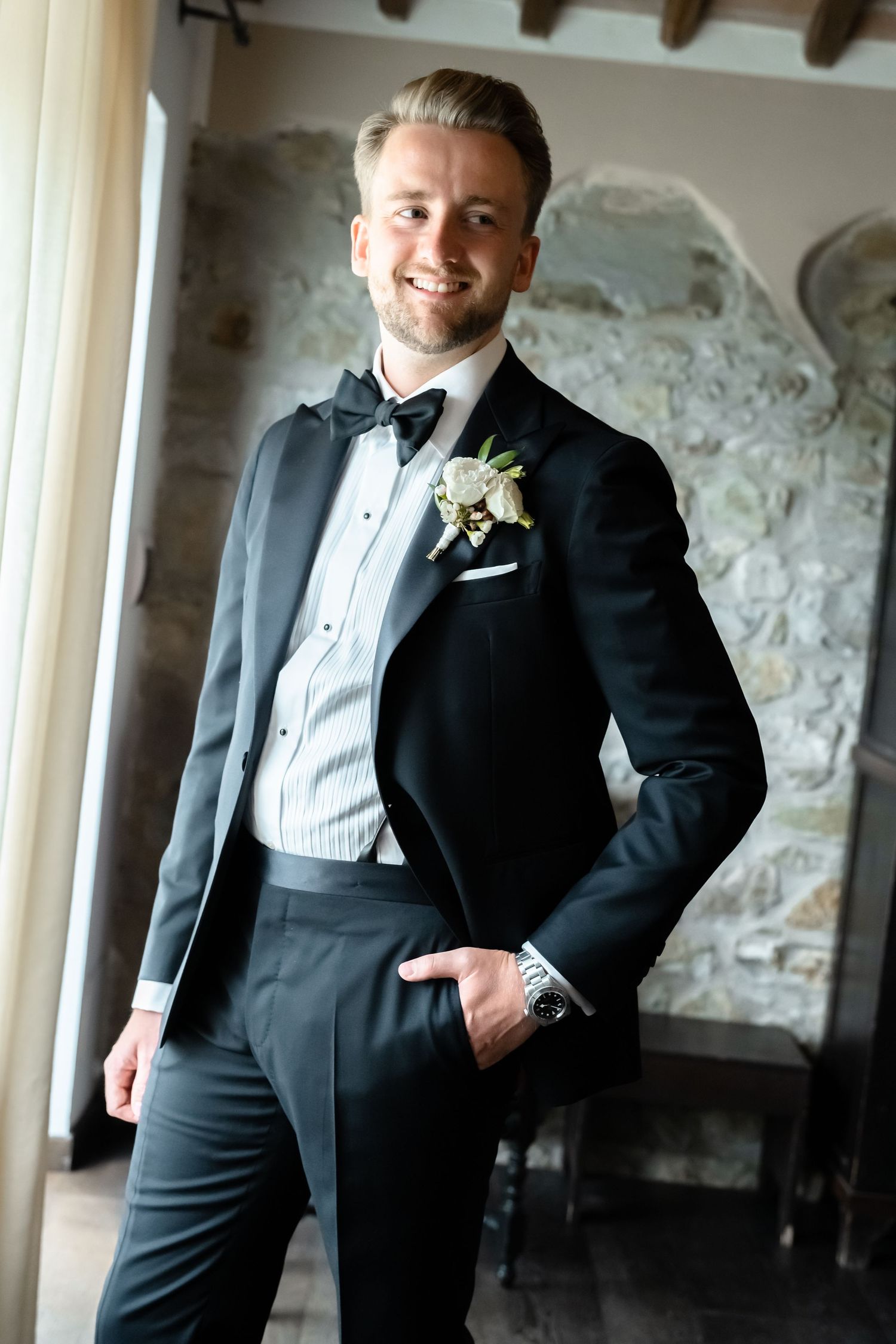 Smiling groom in black tuxedo with boutonniere and bow tie, hands in pockets, posing in elegant stone-walled venue.