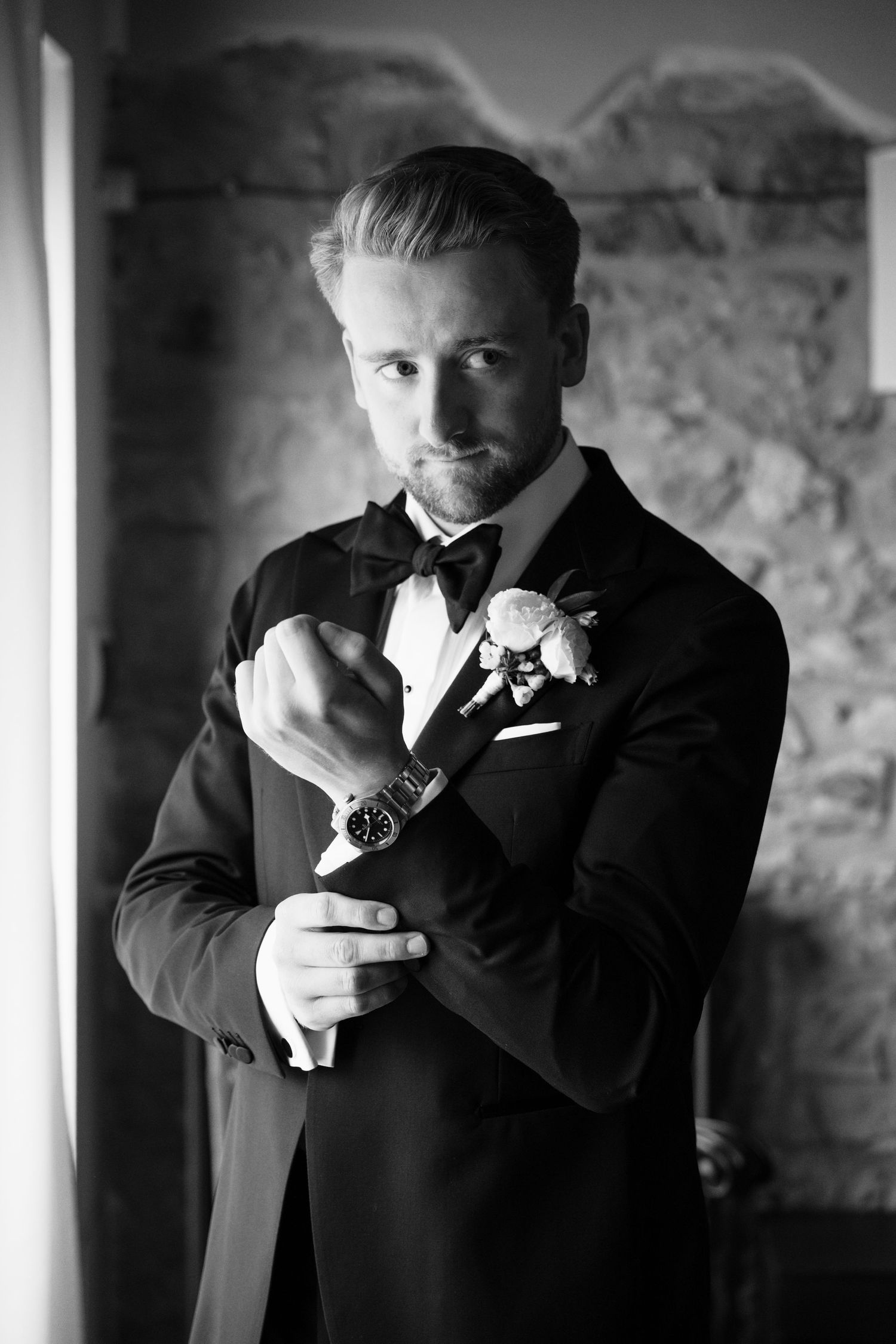 Black and white portrait of a groom in a tuxedo with boutonniere adjusting his watch cufflinks before wedding.