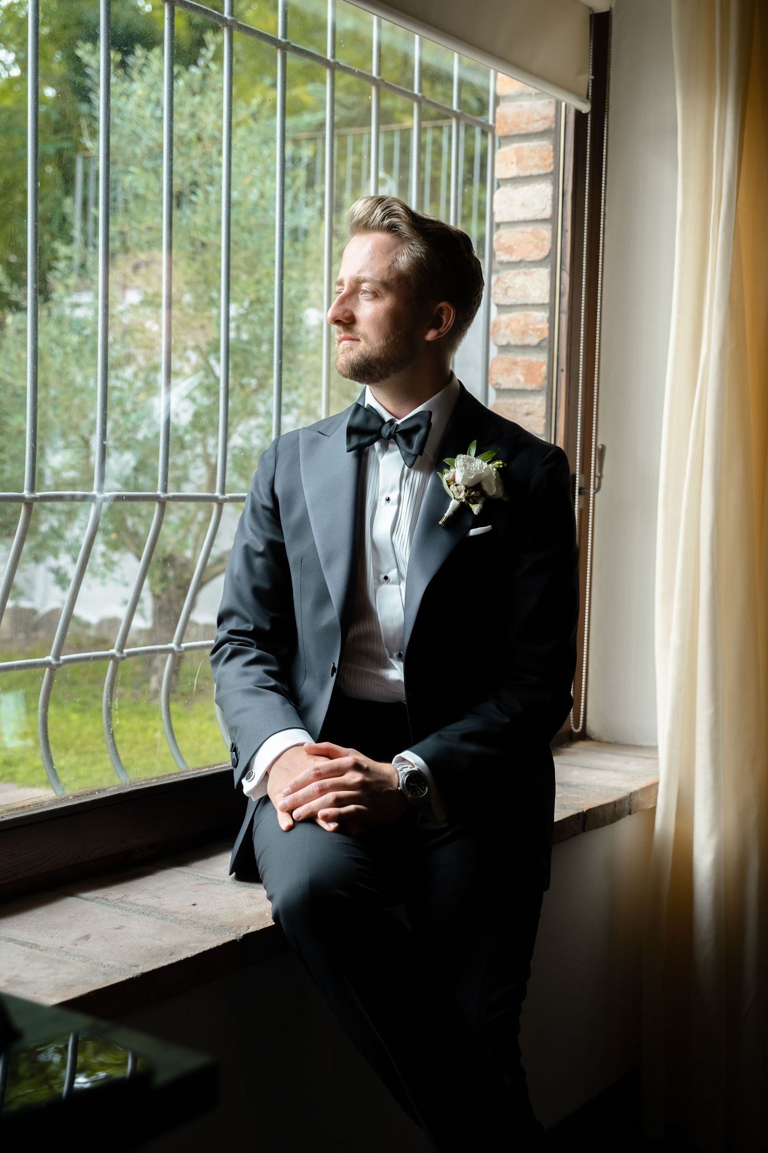 Groom in black tuxedo with bow tie and boutonniere sits on windowsill, gazing thoughtfully outside on wedding day.
