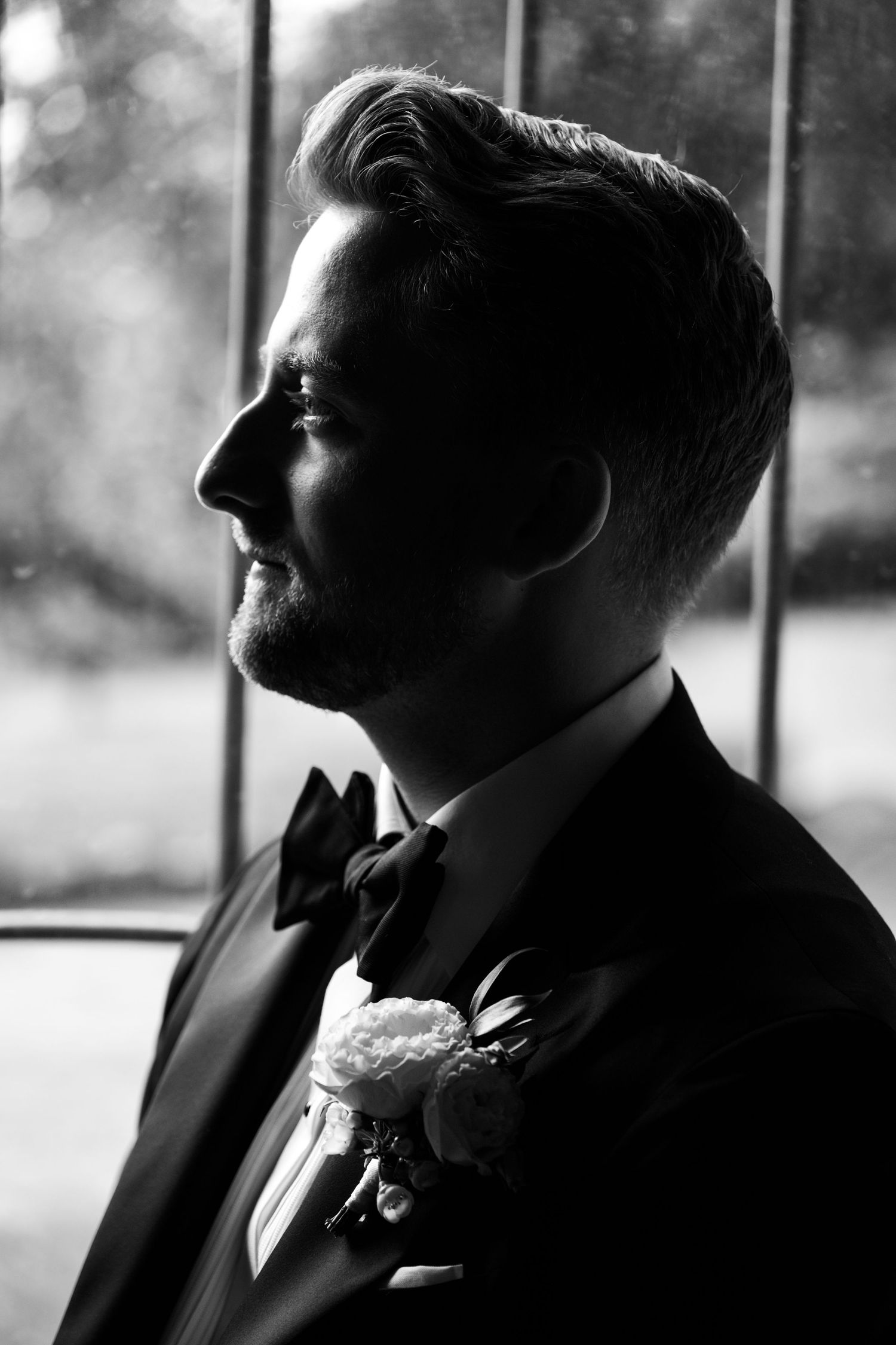 Black and white portrait of a groom in a tuxedo with boutonniere and bow tie, gazing thoughtfully by a window.