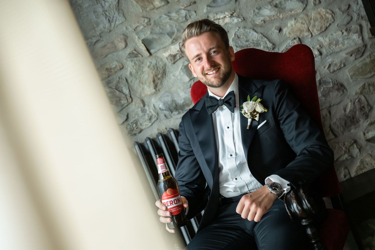 Groom in black tuxedo sitting in red chair holding beer bottle, smiling, with boutonniere and watch on wedding day.
