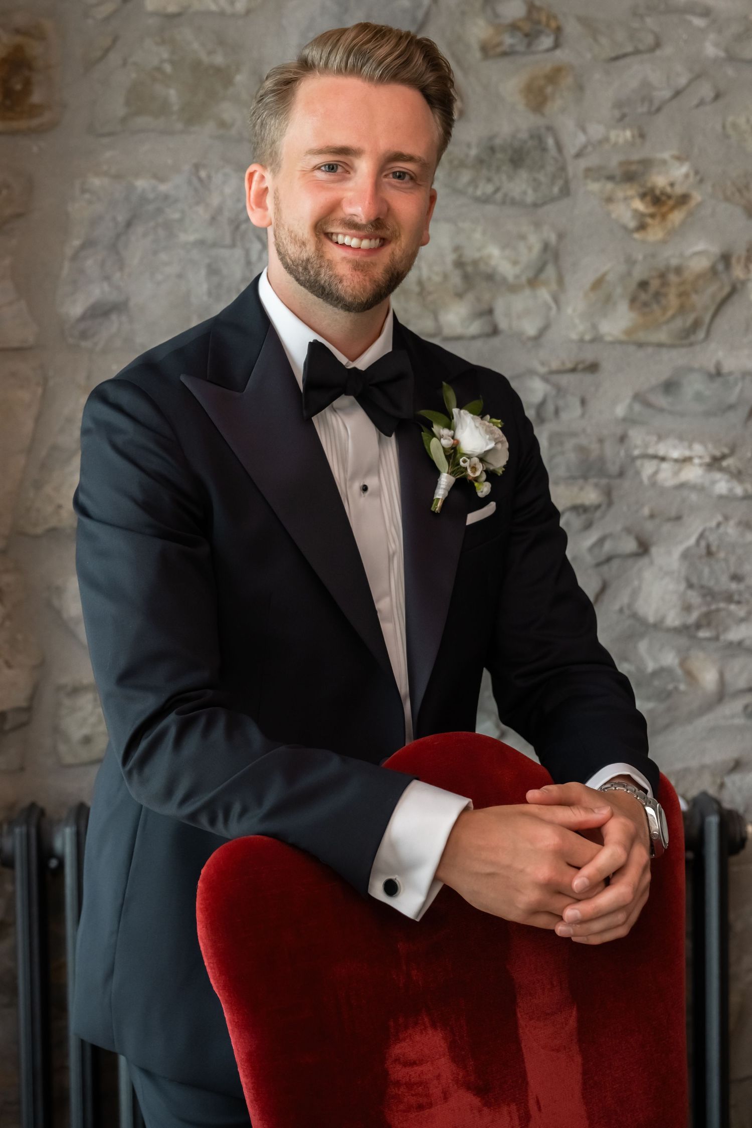 Smiling groom in black tuxedo with white boutonniere leans on red velvet chair against stone wall.