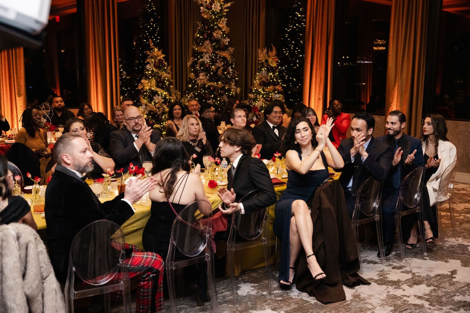 Elegantly dressed guests applaud at a festive holiday gala dinner with Christmas trees glowing in the background.