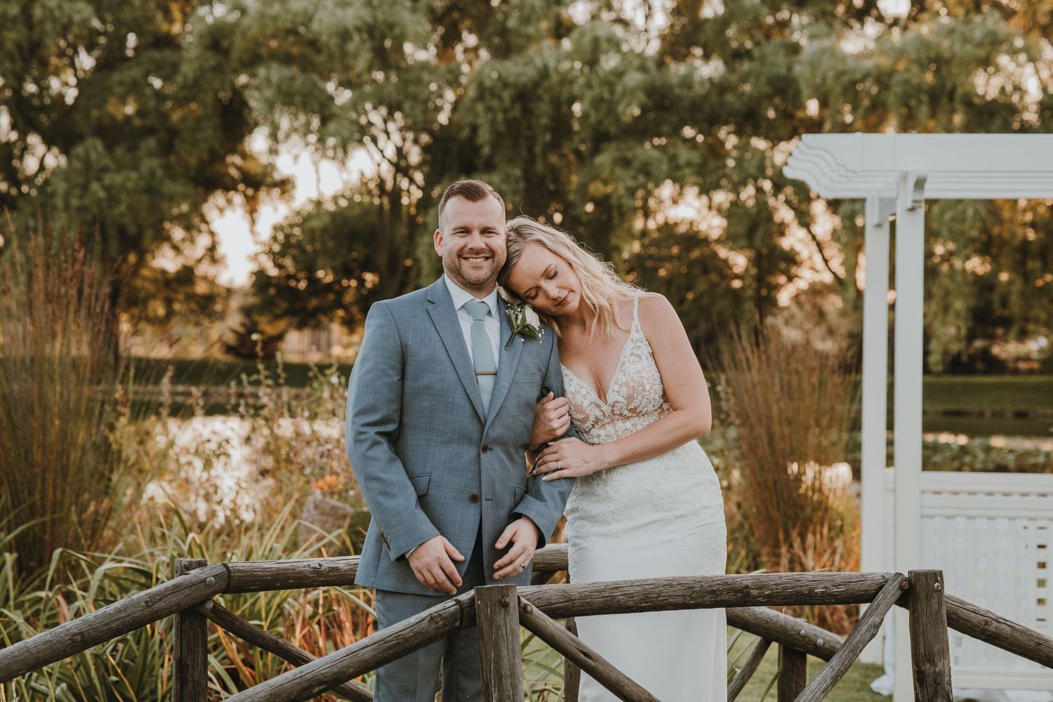 A happy couple enjoying their wedding day on a wooden bridge by a pond at sunset.
