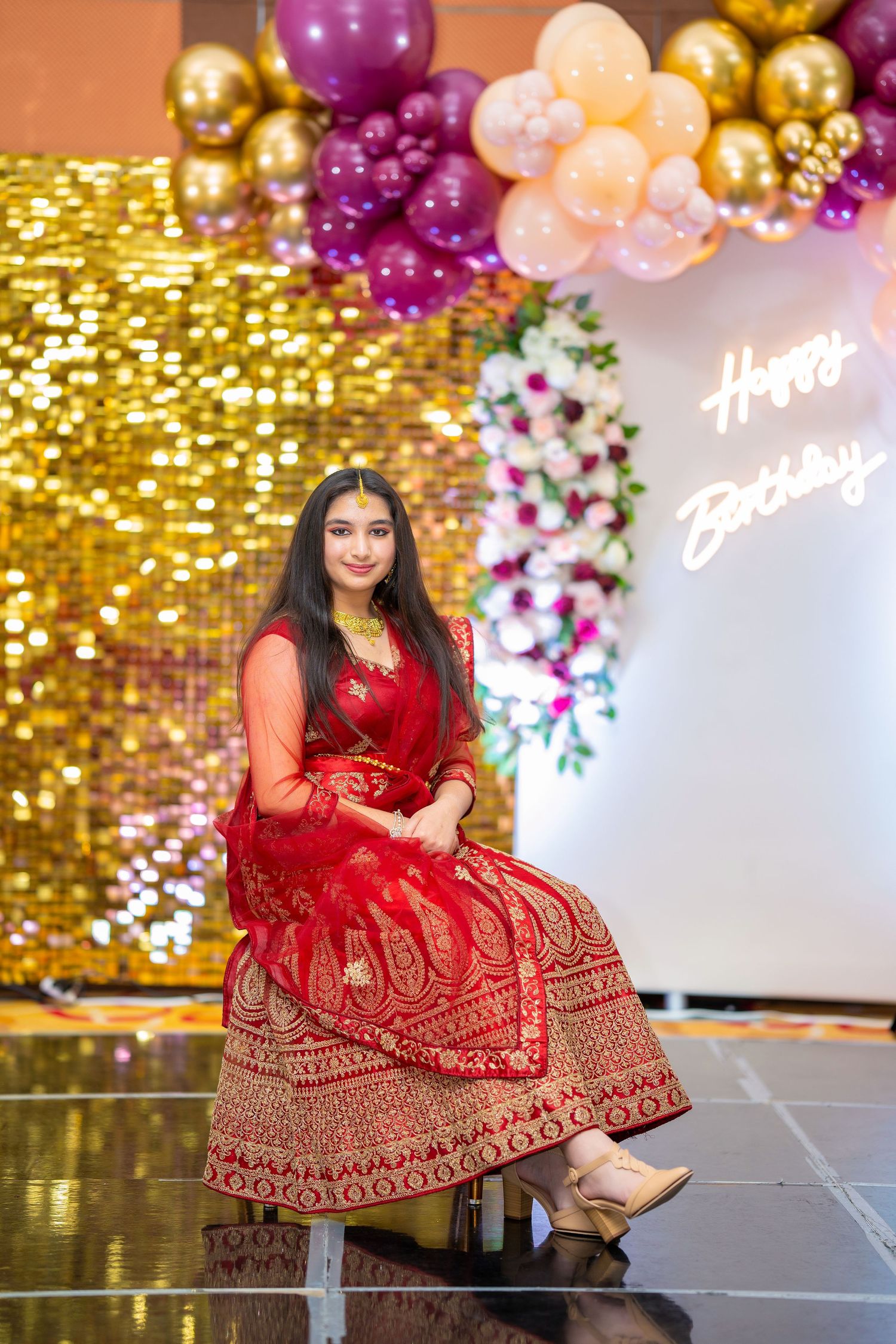 A person in a red traditional dress poses against a backdrop with gold sequins and pink balloons.