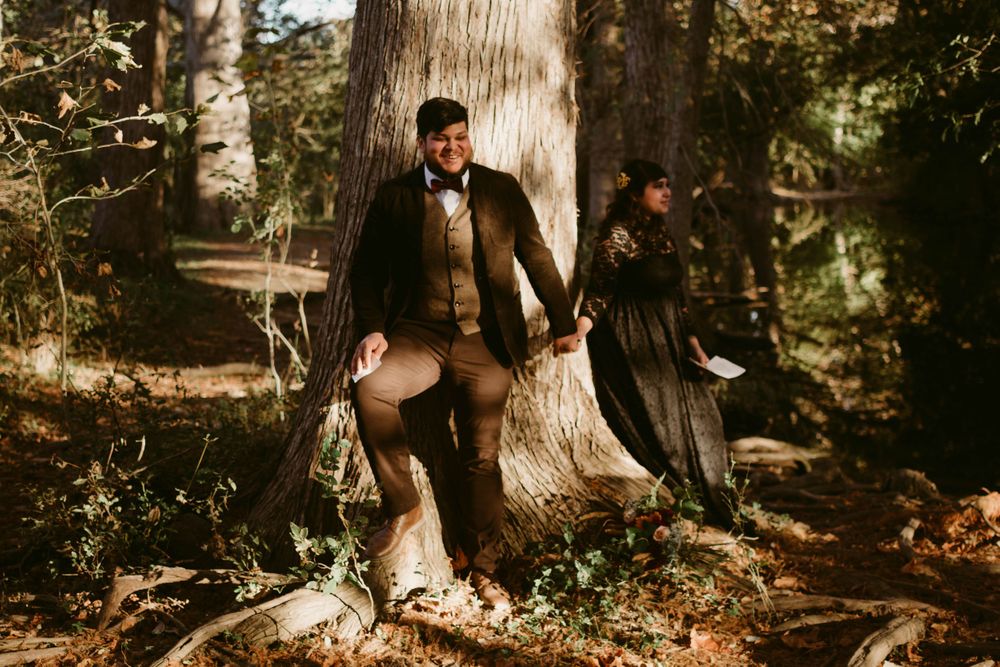 Couple in vintage clothing poses dramatically against tree in wooded setting.