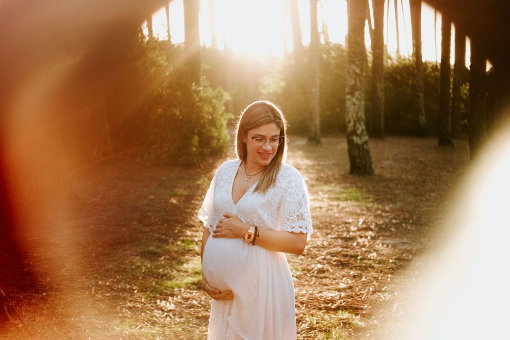 Pregnant woman in white lace dress cradling her baby bump in a sunlit forest at golden hour.