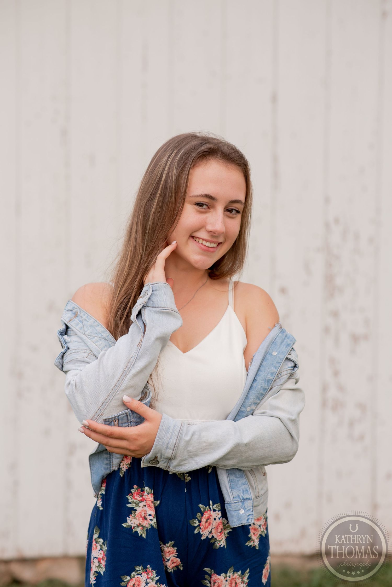 Casual portrait wearing white tank top and denim jacket against rustic white wall background.