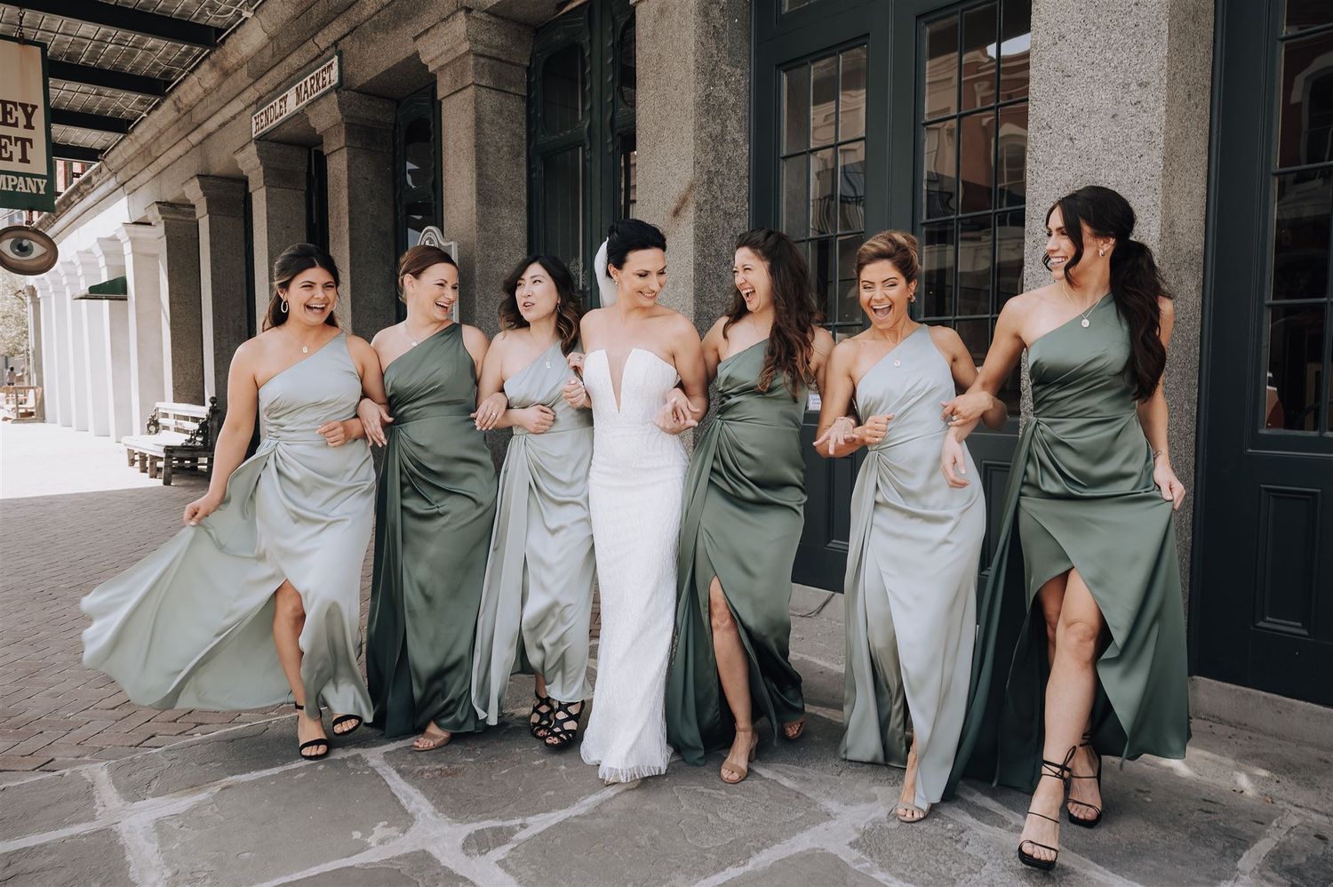 Bridal party in sage green dresses poses on historic street.