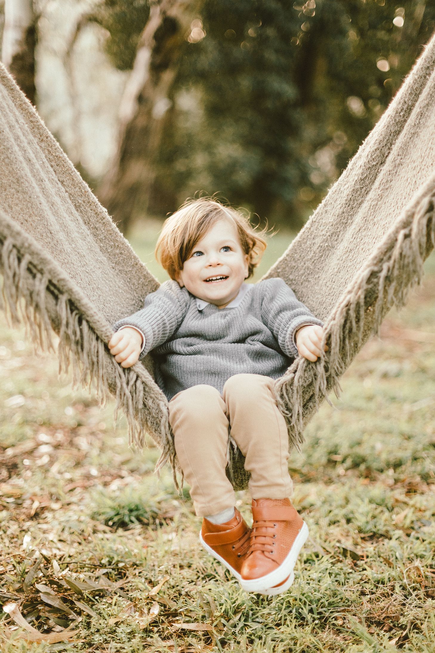 Small child in a gray sweater playing on a rustic rope swing in an outdoor setting.