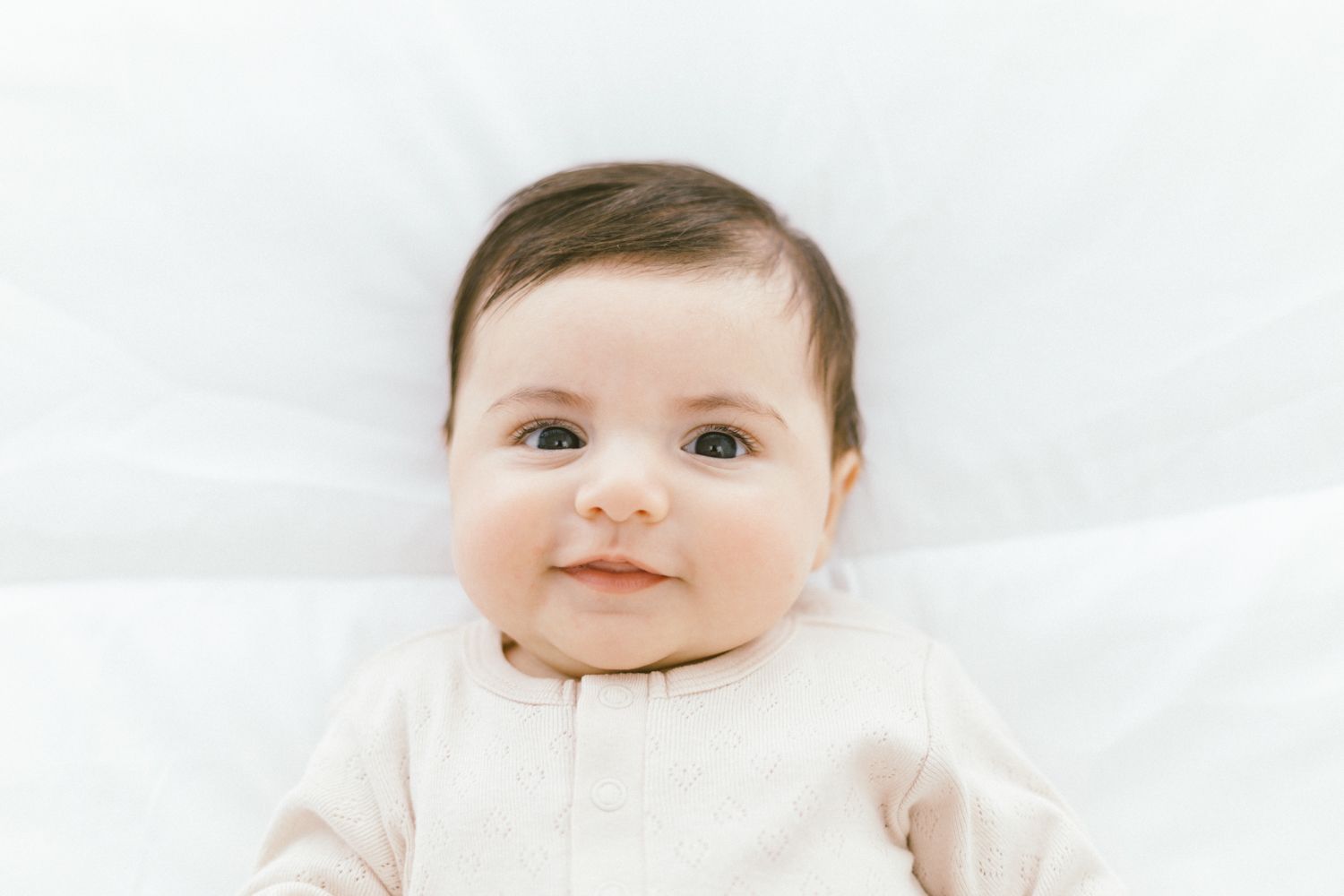 Baby wearing cream colored outfit lying on white bedding with bright smile.
