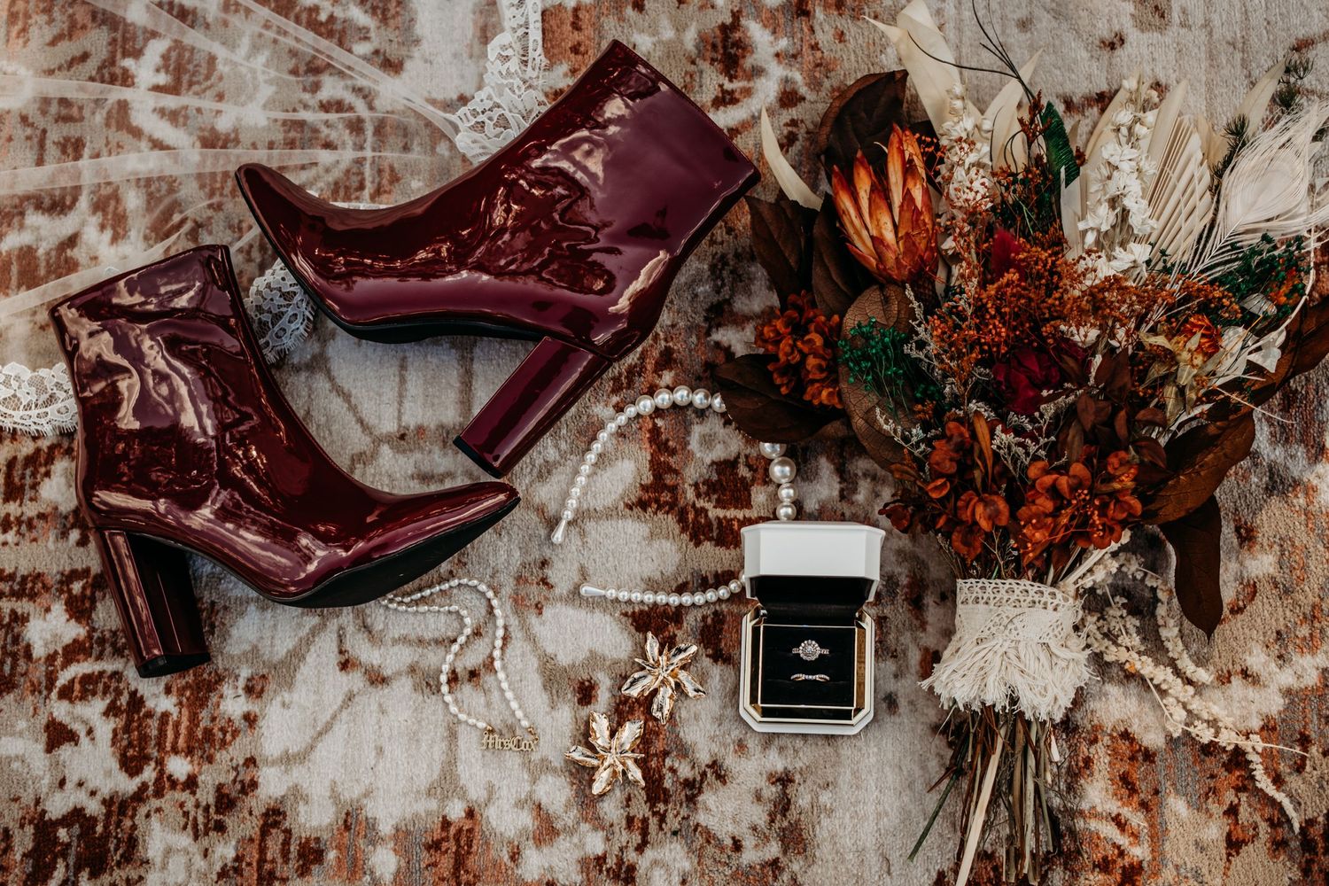 Burgundy leather wedding accessories and floral arrangements styled against a textured surface.