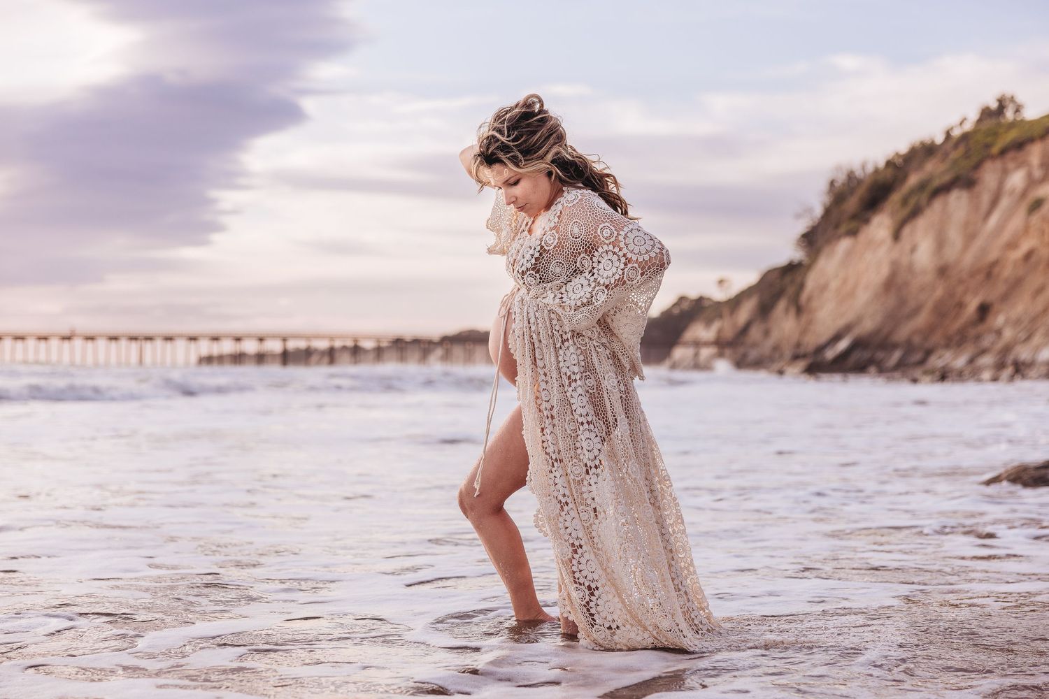 Ethereal white lace dress catches the wind on a scenic beach with cliffs and pier.