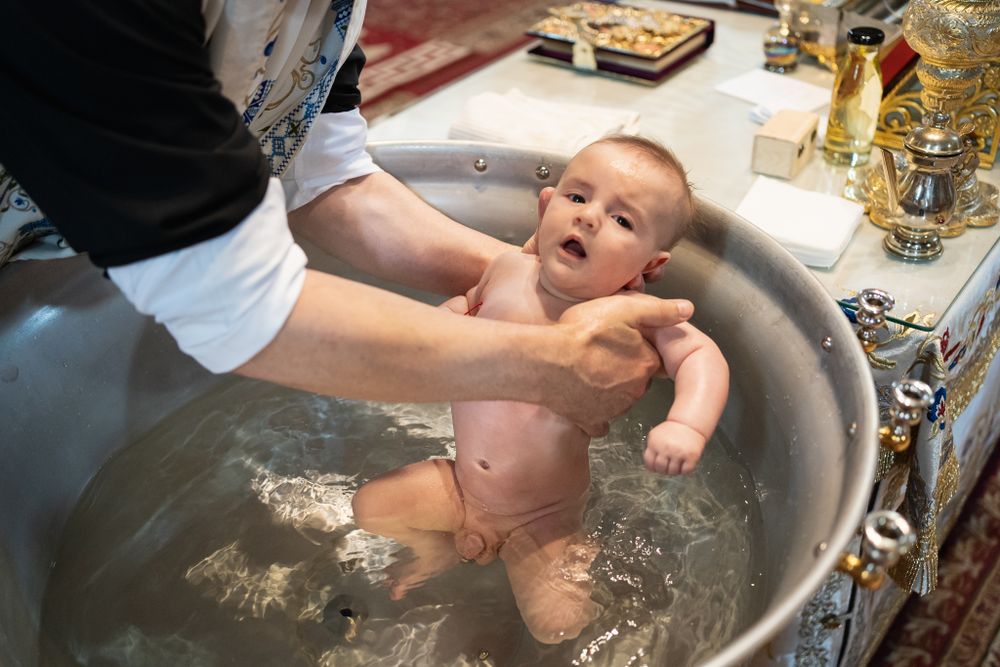 Infant baptism ritual taking place in a metal basin with holy water.