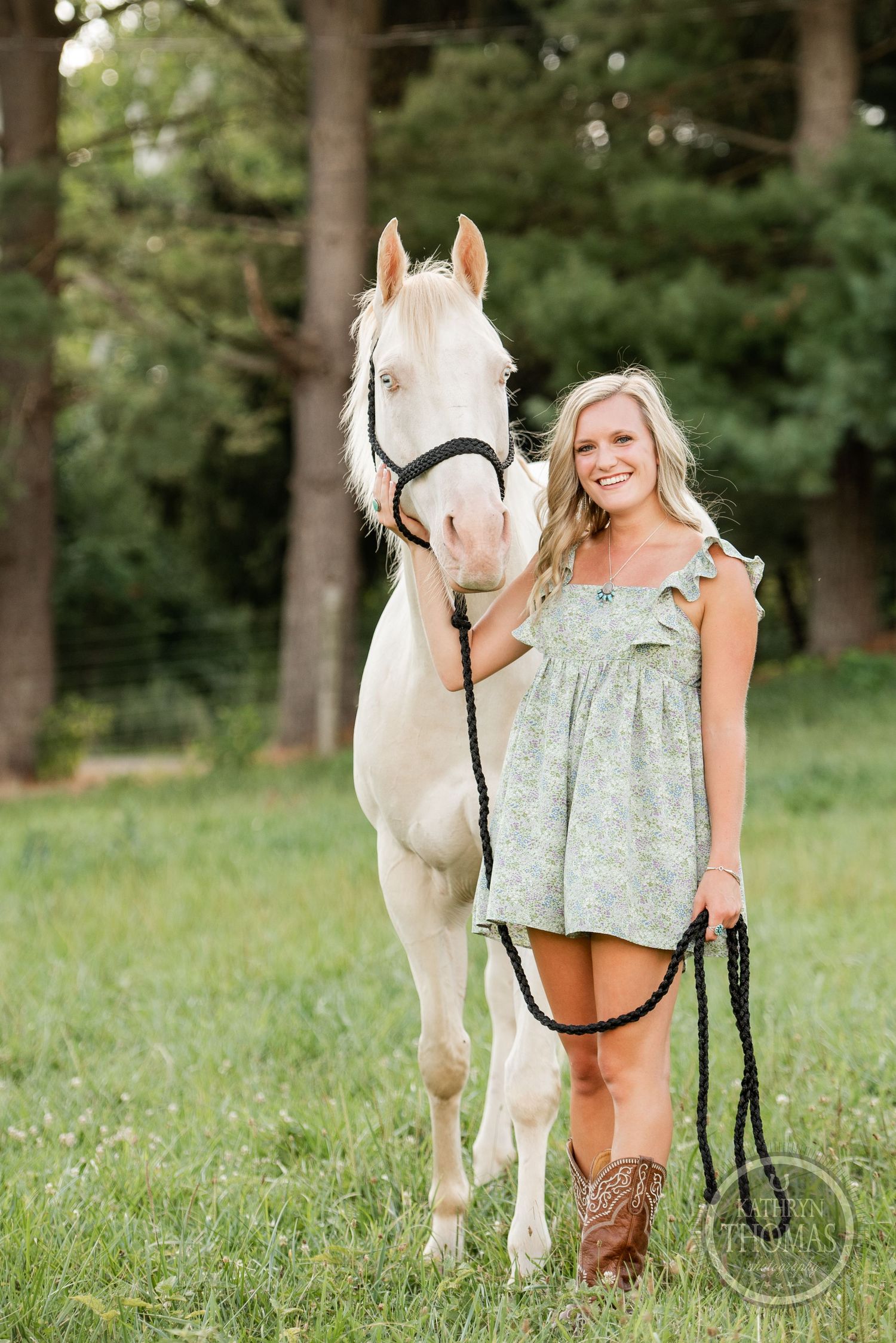 A beautiful white horse stands with its owner in a sage dress and cowboy boots against a wooded backdrop.