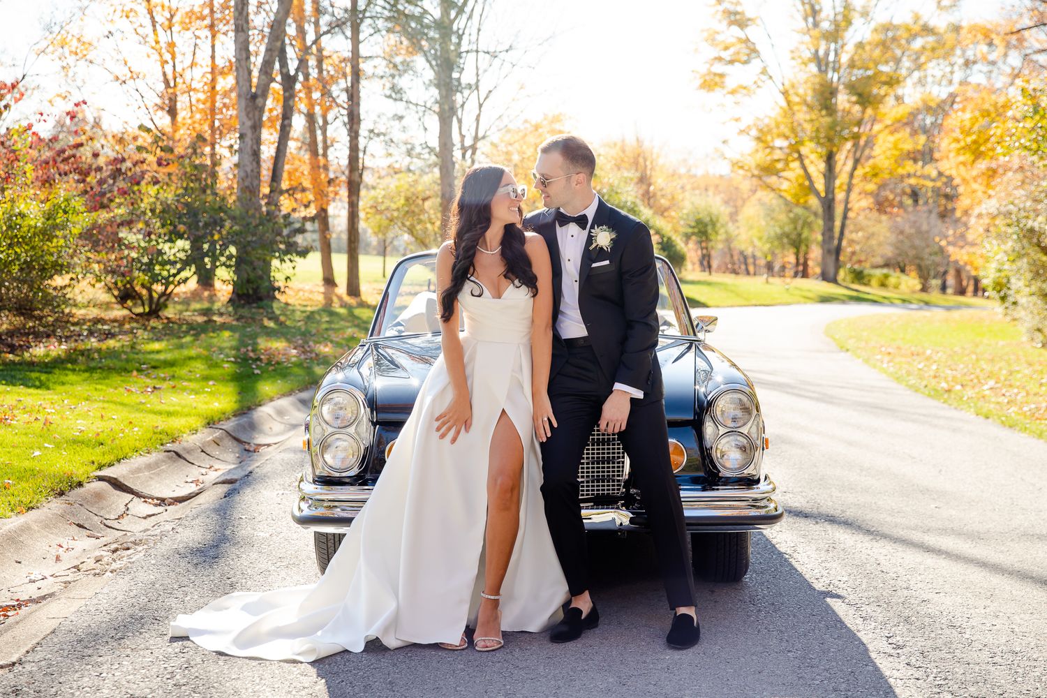 Newlyweds pose together beside vintage car on autumn tree-lined road.