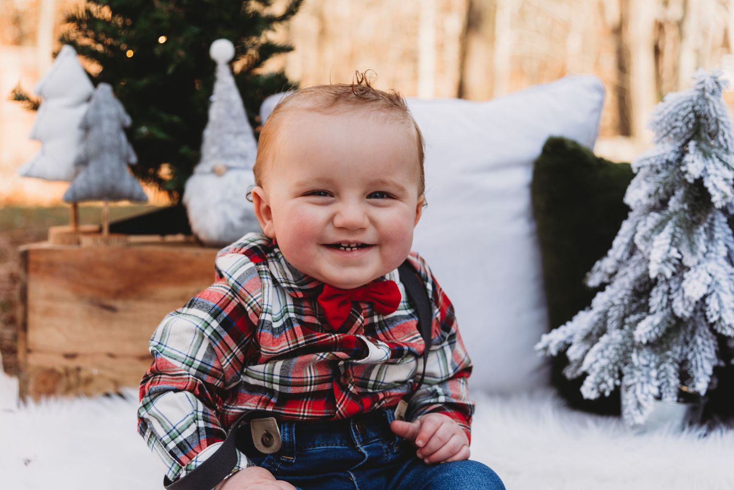 A smiling toddler in a plaid shirt and jeans sits among winter holiday decorations.