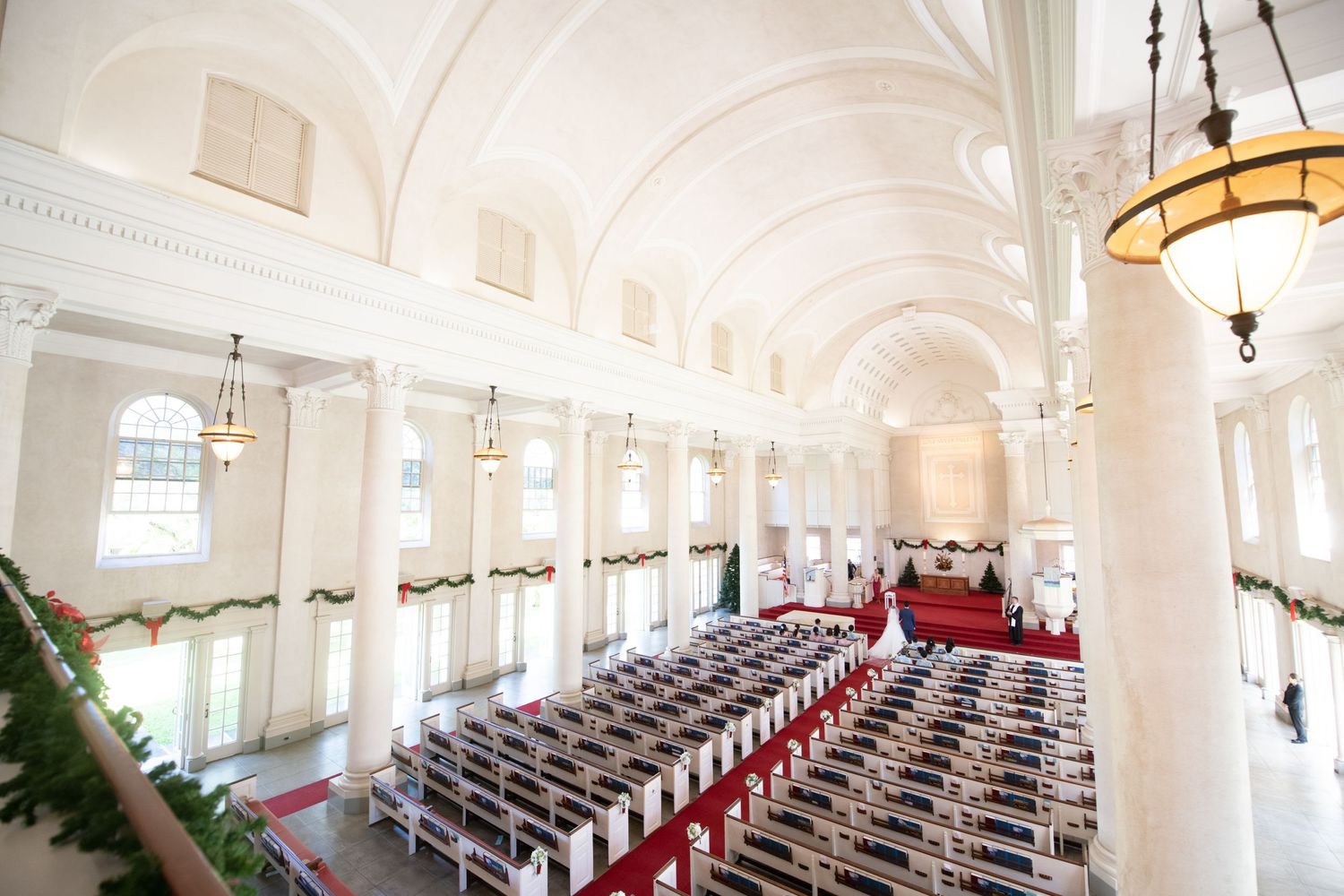 Grand white church interior with arched ceilings, rows of pews, and holiday garland decorations viewed from above.
