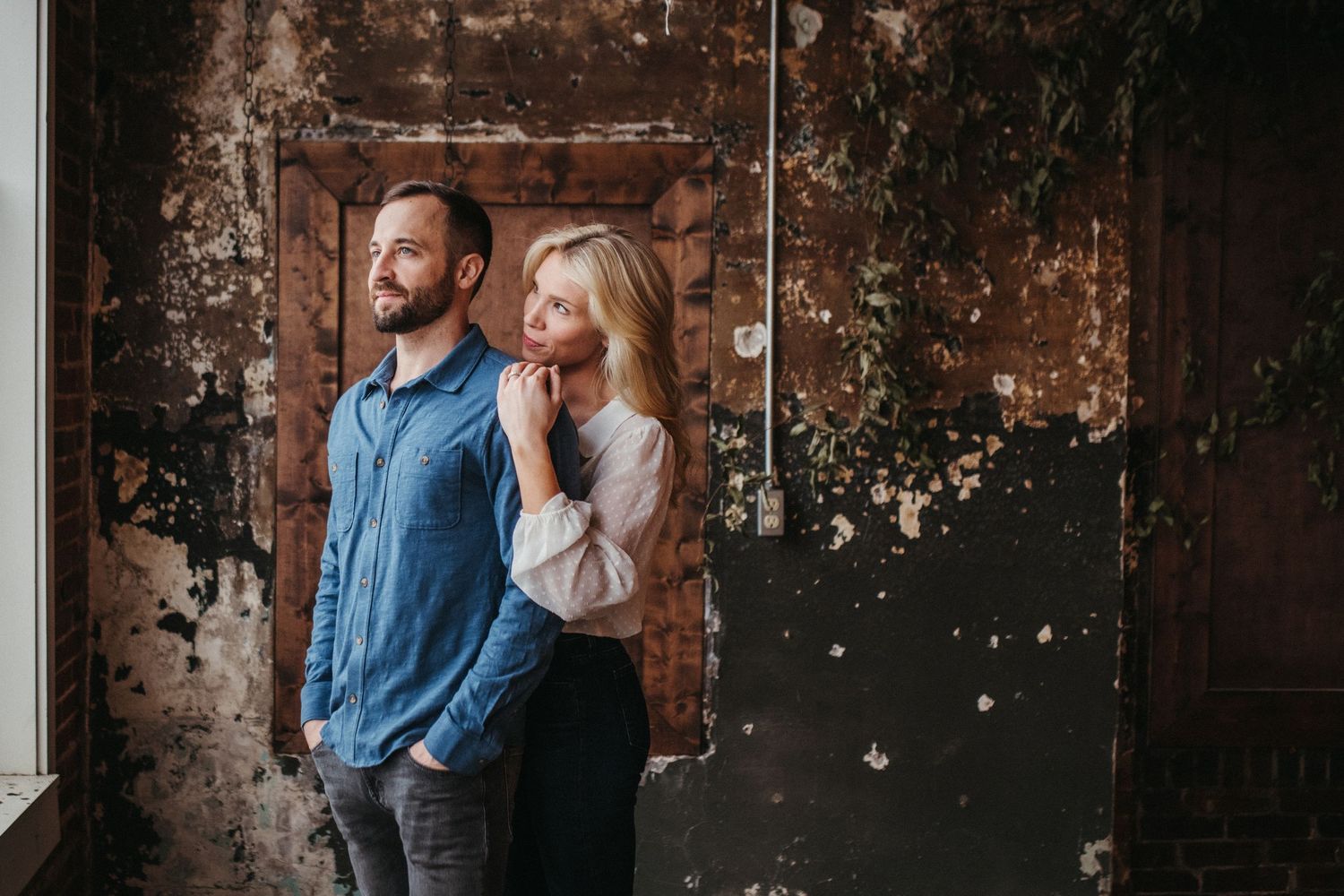 A couple poses against a rustic weathered wall in casual attire.