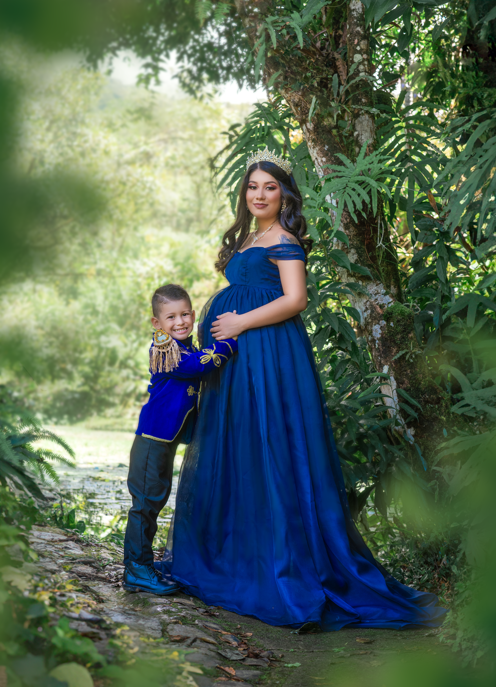 Maternity photo of mother and child in matching royal blue outfits against lush greenery.