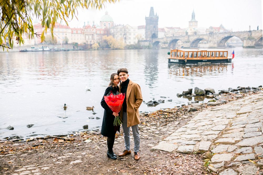Couple shares a romantic moment by Prague's Vltava River with tourist boat in the background.