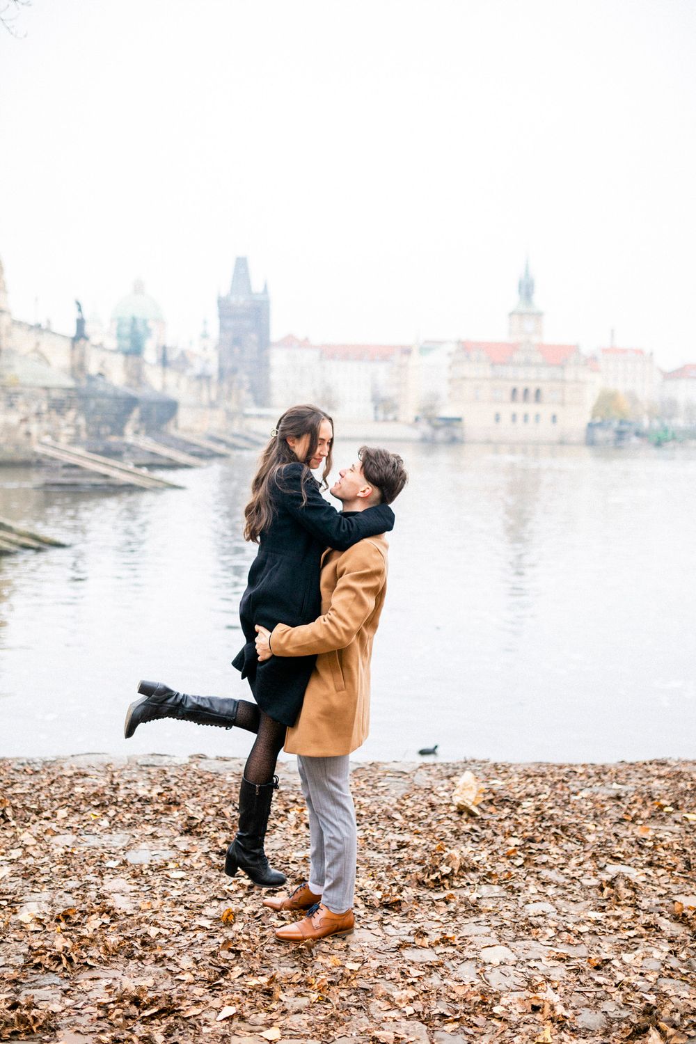 Romantic couple embracing on banks of Charles River in Prague with iconic architecture in background.