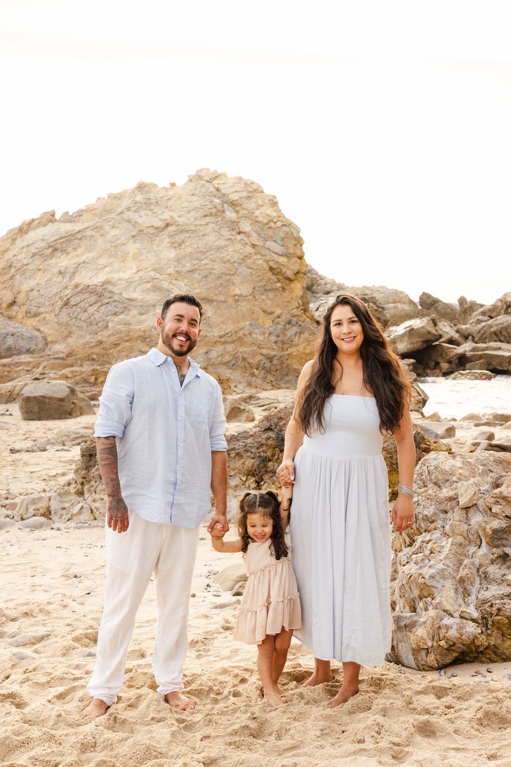 A happy family of three poses on a sandy beach with rocky cliffs, parents holding their young daughter's hands.