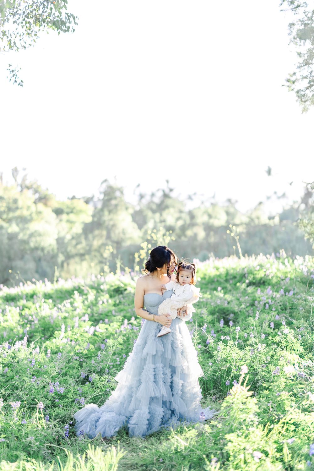 Woman in blue ruffled gown holds toddler with floral crown in a sunlit wildflower meadow.