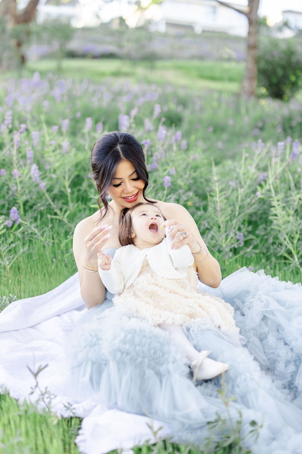 A smiling mother in a blue tulle gown holds her laughing baby girl in a lavender field during an outdoor photo session.