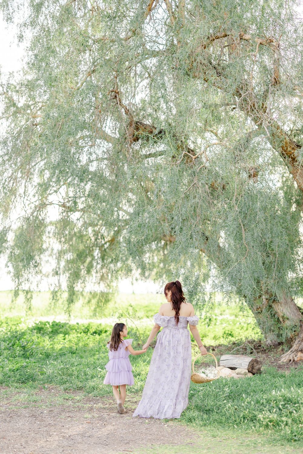 A mother and daughter in matching lavender dresses hold hands under a weeping willow tree in a lush green field.