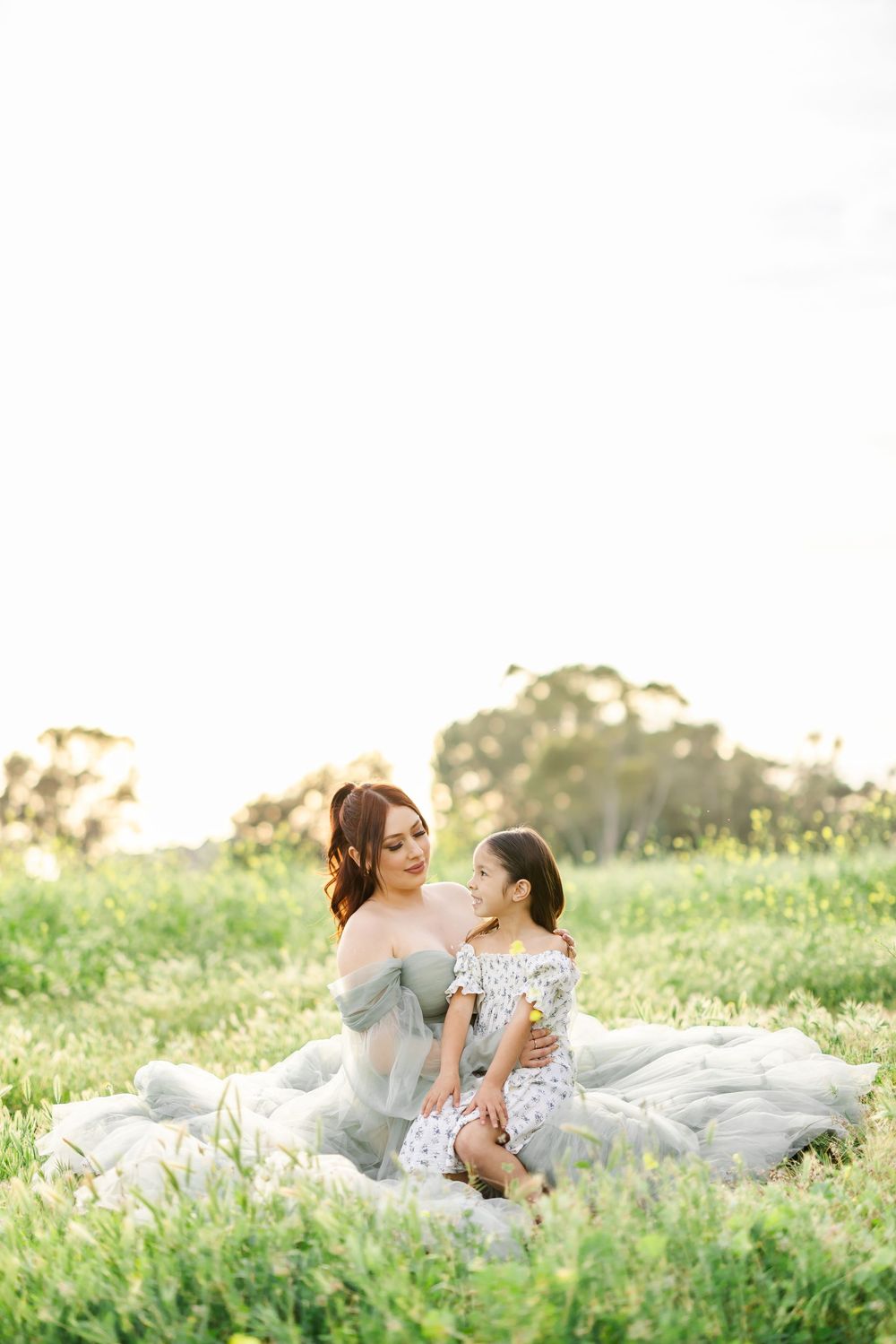 Mother and daughter sitting together in a sunlit green meadow, sharing a tender moment during an outdoor photo session.