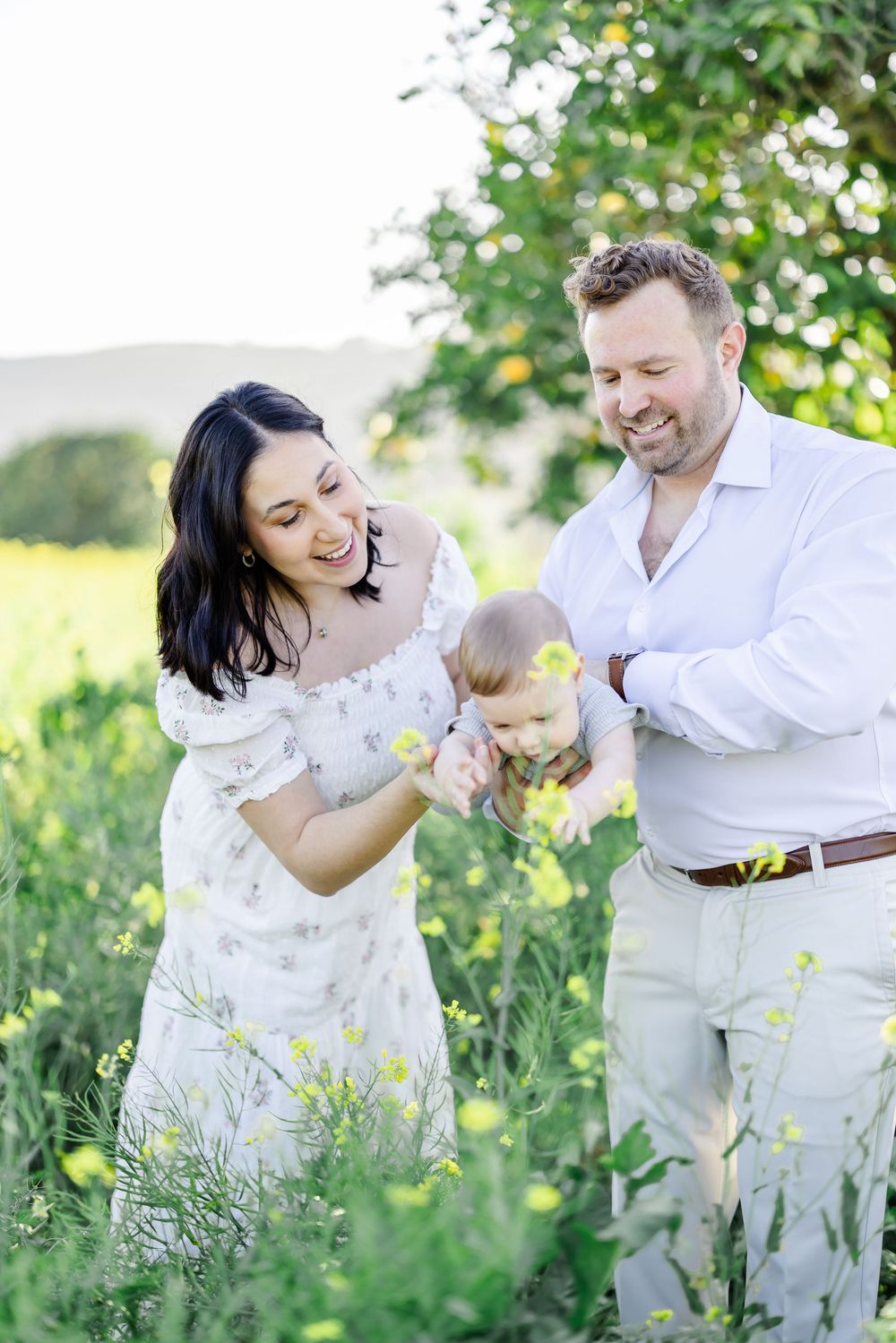 Happy family with baby enjoying a sunny day in a yellow wildflower field, parents smiling at their infant child.