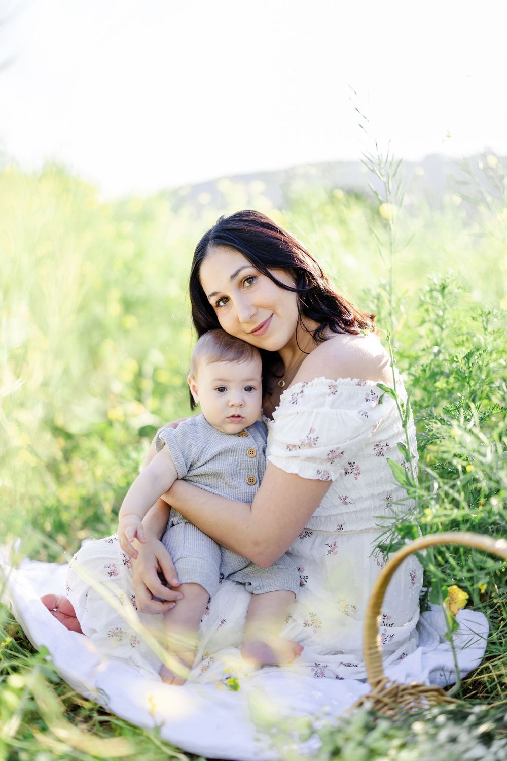 A smiling dark-haired mother in a floral dress holds her baby outdoors in a sunny green meadow with a wicker basket nearby.