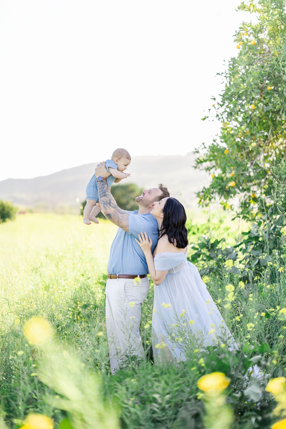 A joyful couple lifts their baby in a sunny green meadow with yellow wildflowers and mountains in the background.