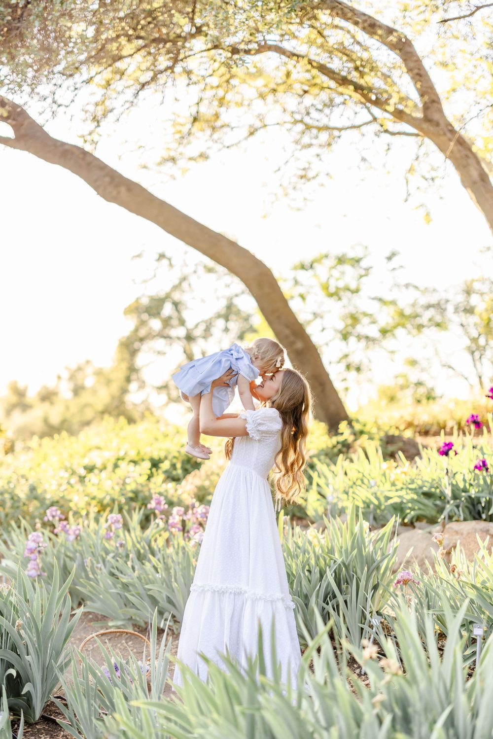 A sunlit garden scene with white dress and baby among iris flowers and dappled light.