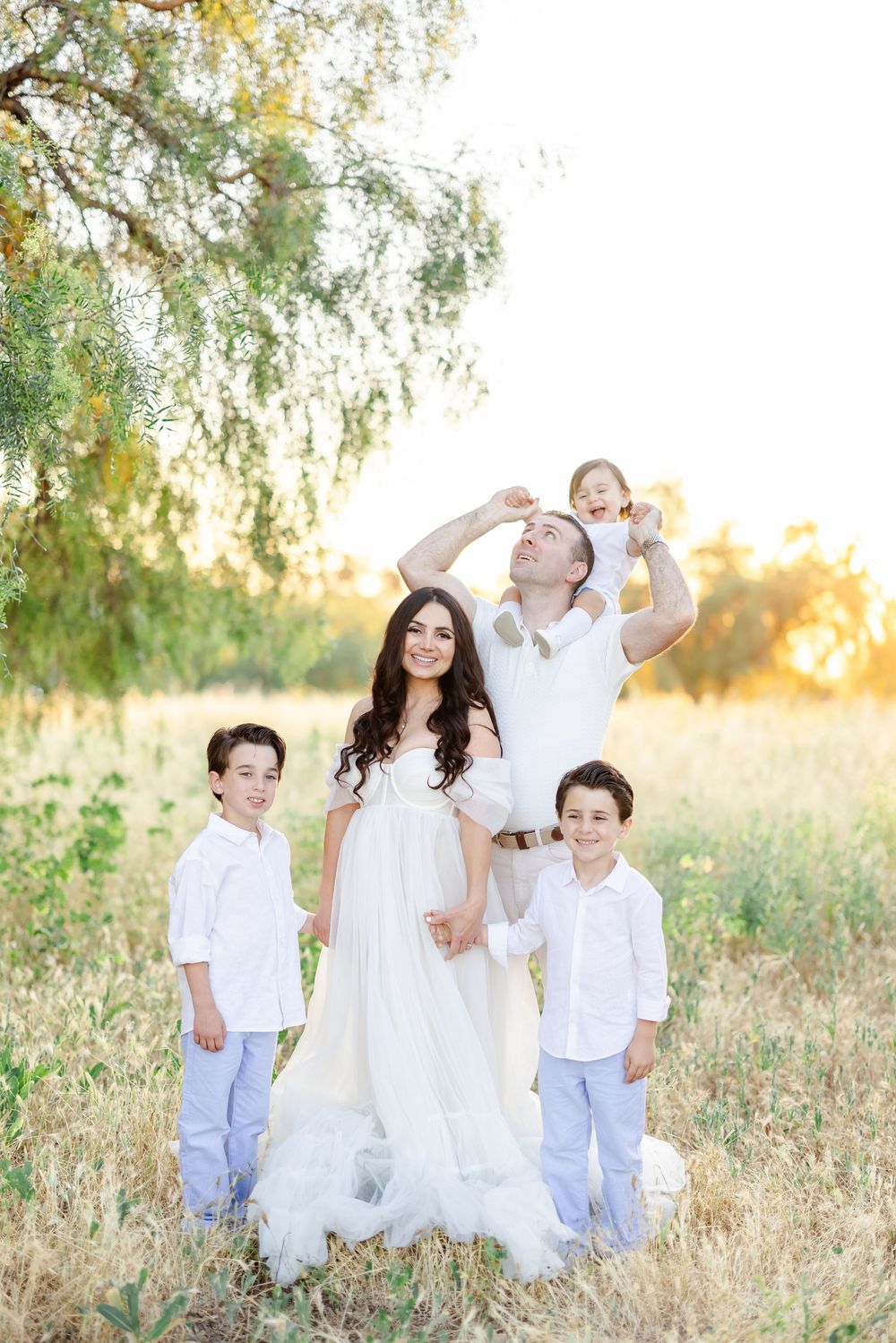 Happy family in white outfits posing outdoors in golden sunlight, parents with three children in a scenic field.
