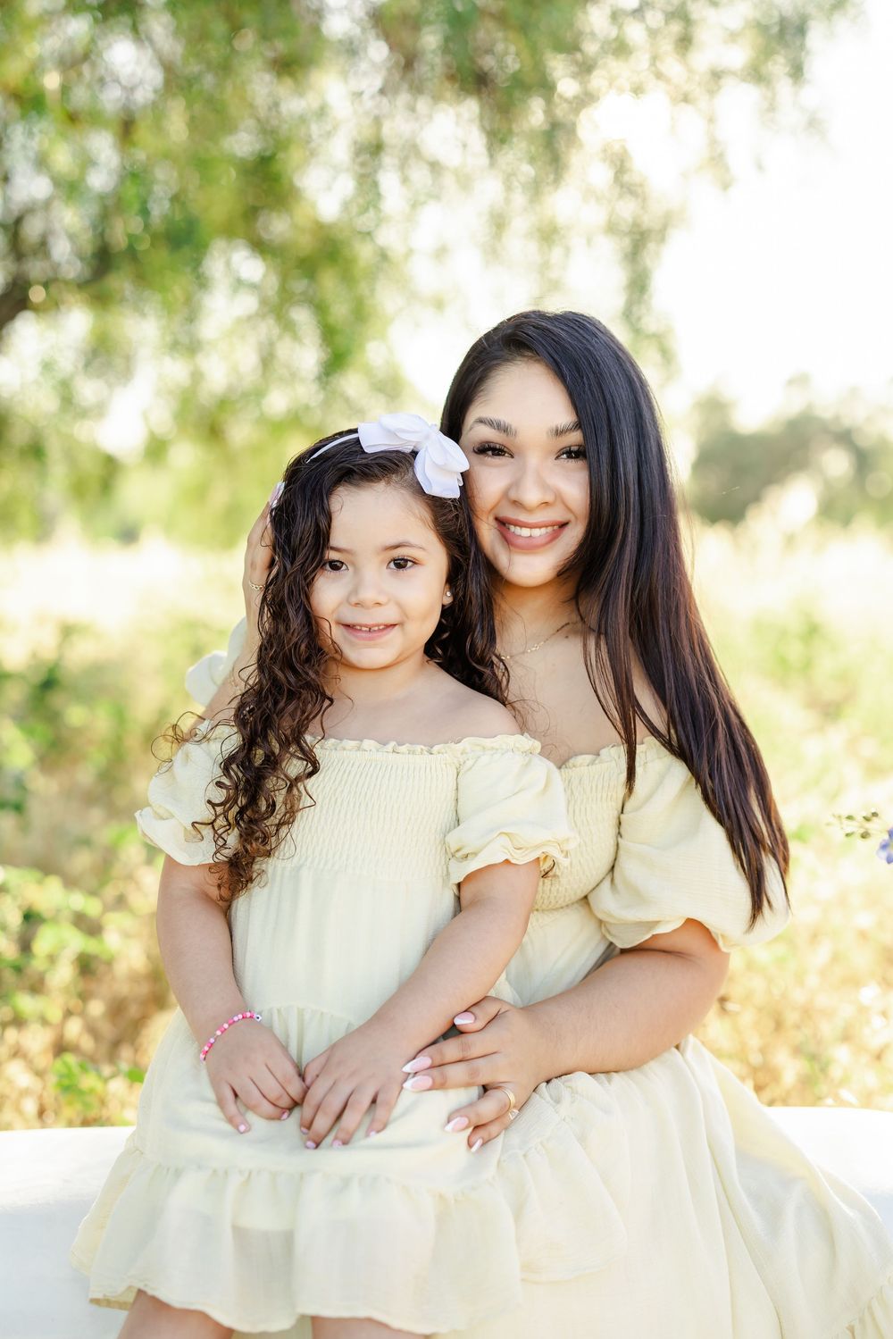 A smiling mother and daughter in matching yellow dresses pose together outdoors surrounded by lush green trees.
