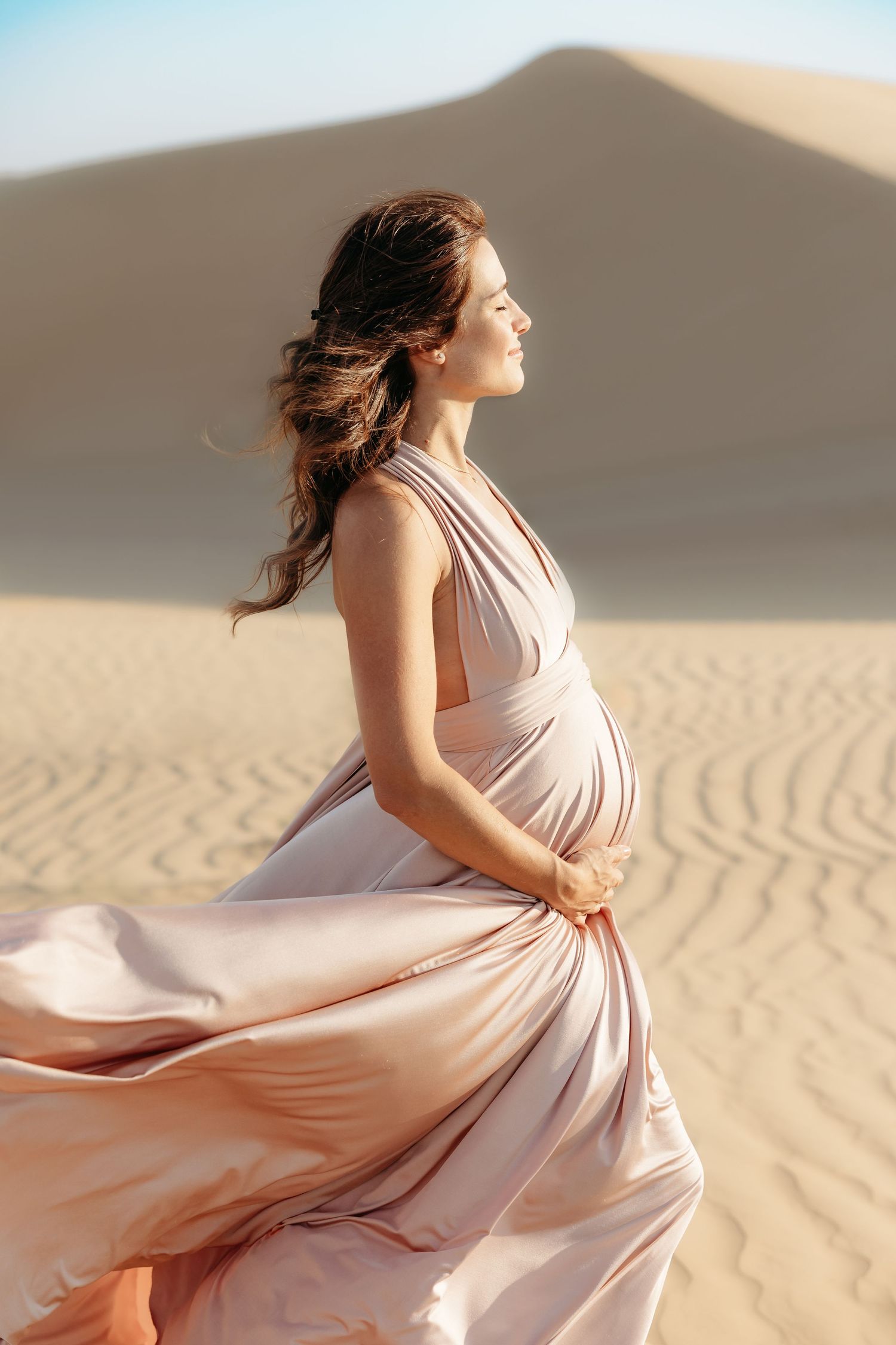 A maternity portrait of a woman in a light pink dress, her gown flowing in the breeze as she stands gracefully against the desert backdrop.