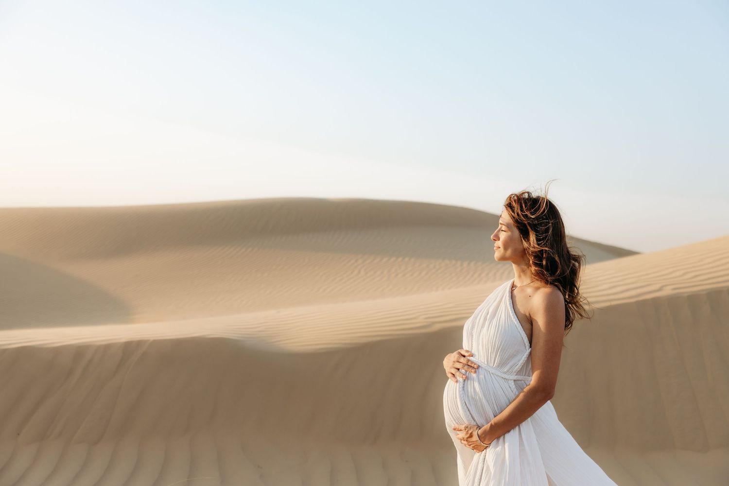 A maternity portrait of a woman in a flowing white dress, standing in the desert and cradling her baby bump, with soft sunlight highlighting