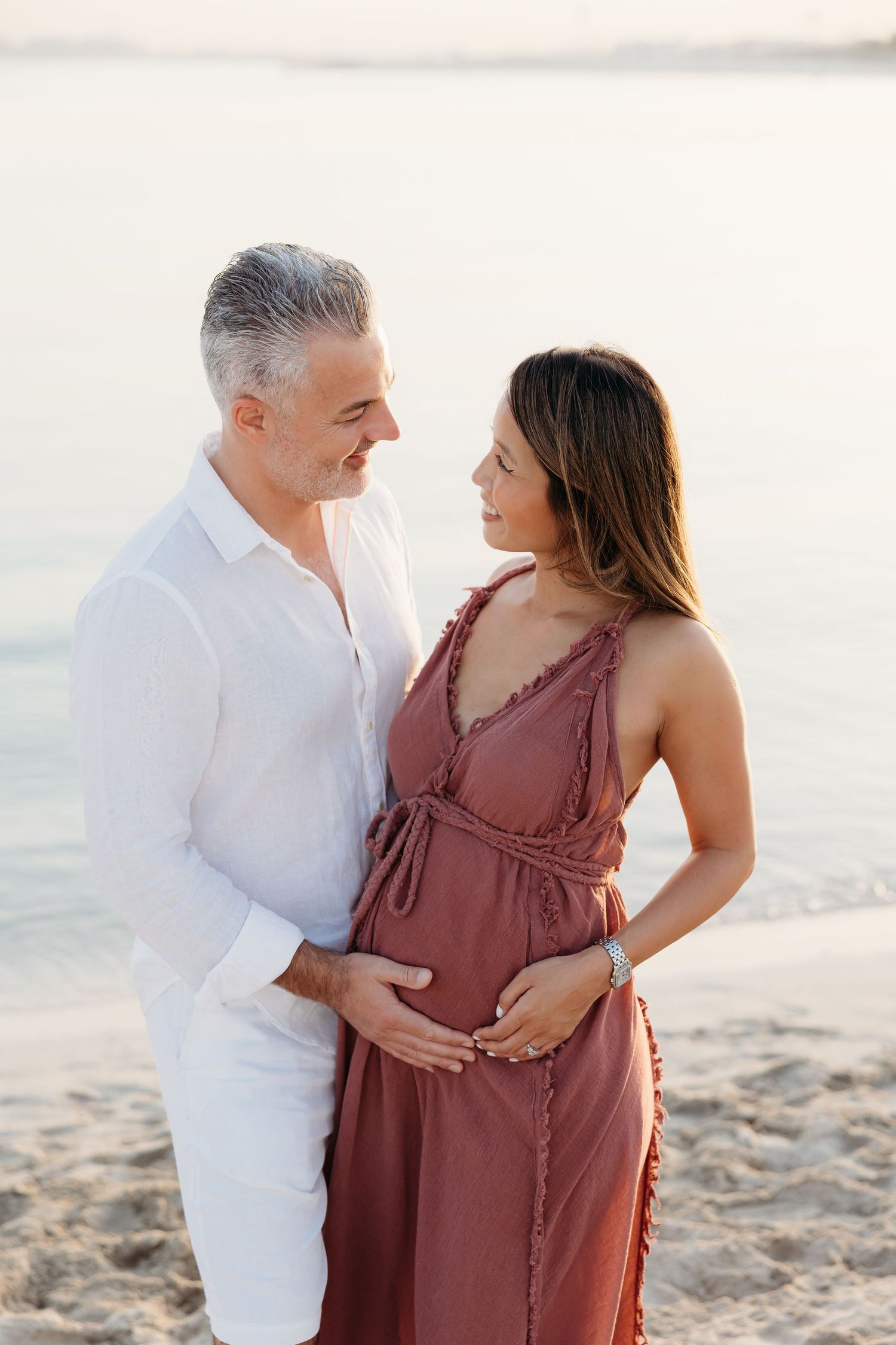A romantic maternity photo of a couple on the beach, with the man in white gently holding the woman’s waist as she cradles her bump.