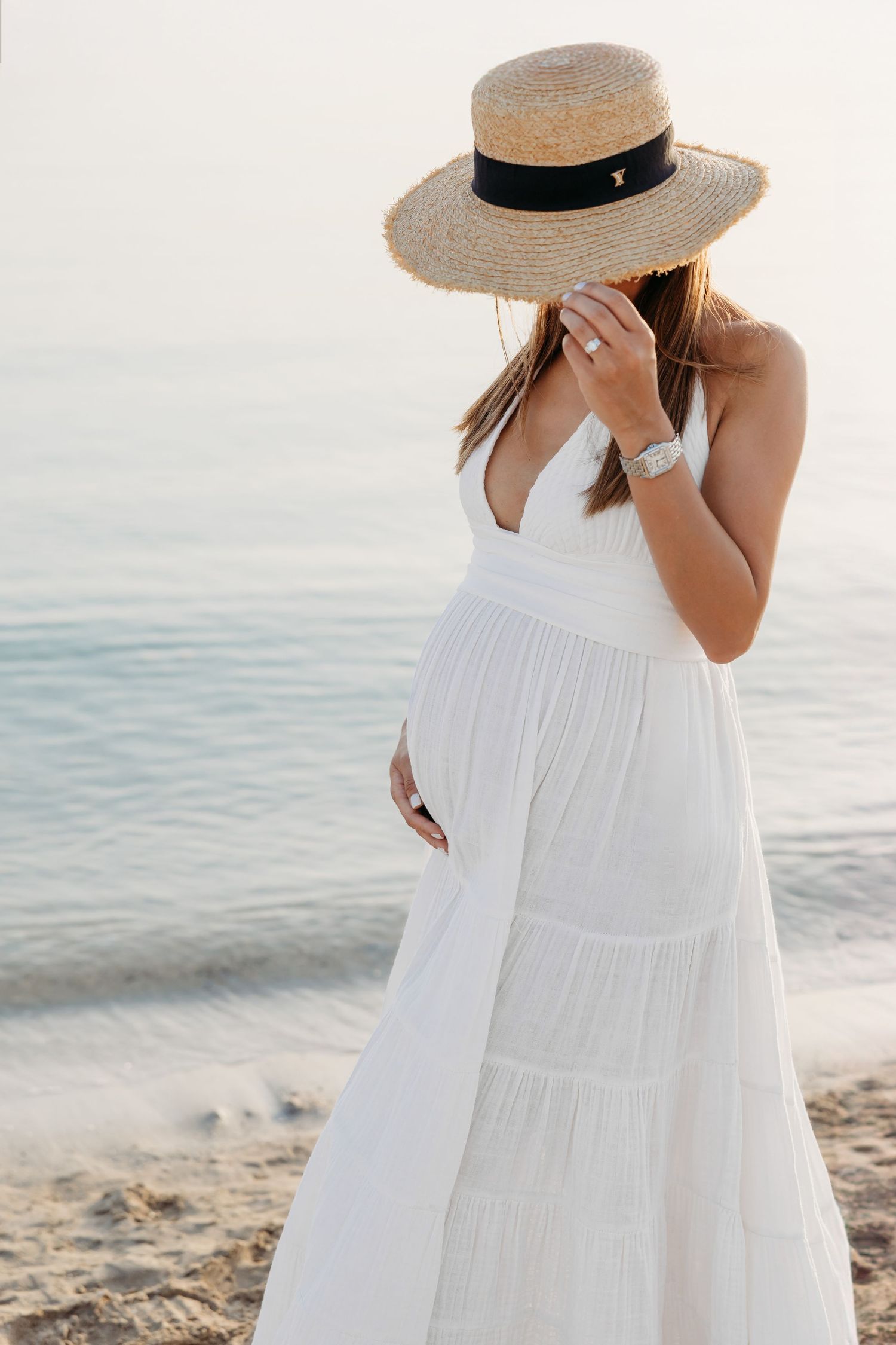 A beach maternity portrait of a woman in a flowing white dress, wearing a wide-brimmed hat, standing gracefully by the shore.