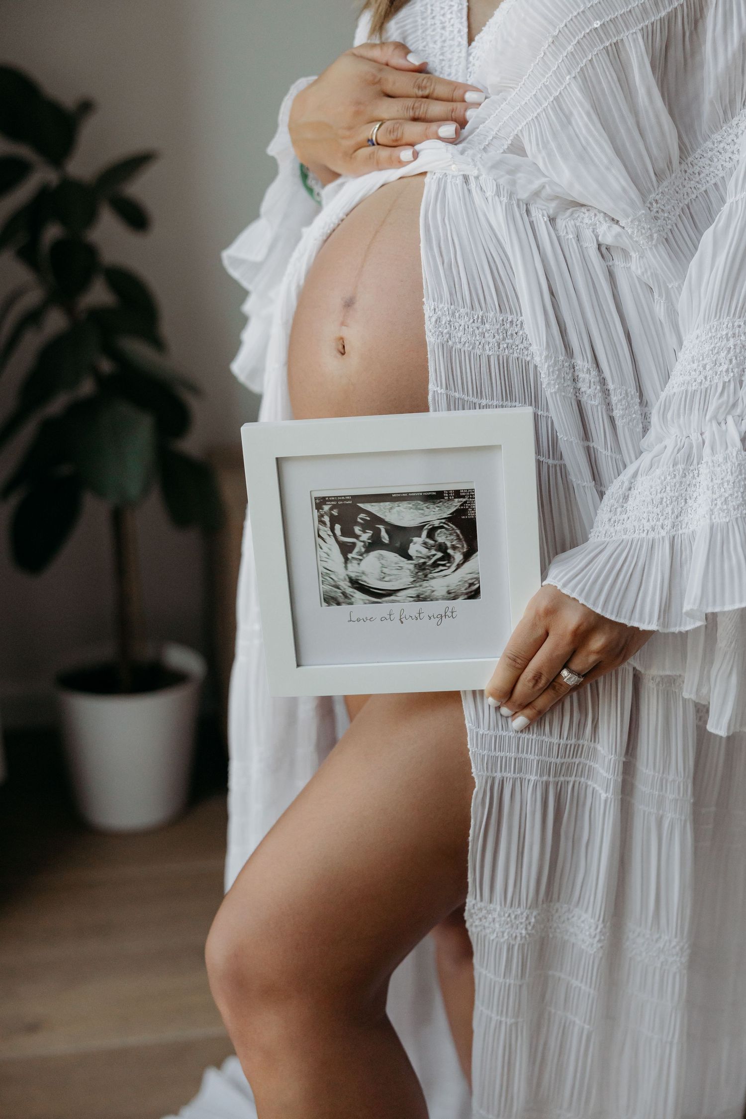 A maternity portrait of a woman in a white robe, holding an ultrasound photo near her baby bump, standing in a softly lit room.