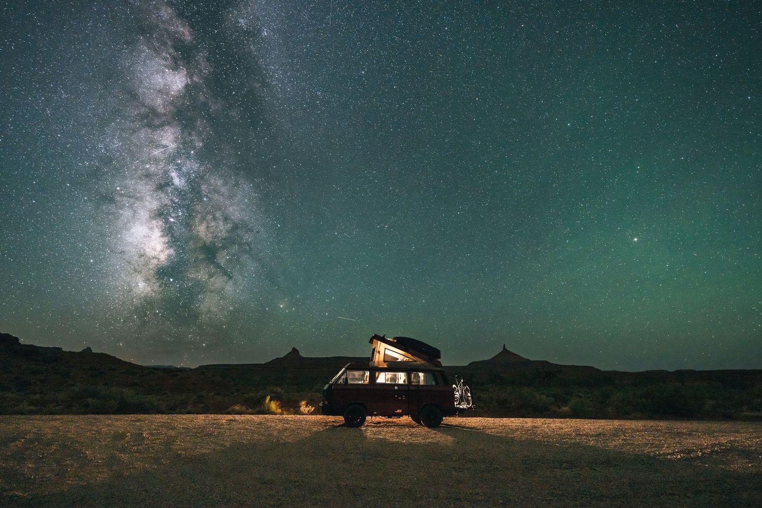 A camping van parked under the Milky Way galaxy with a teal-colored night sky and campfire glow in the foreground.