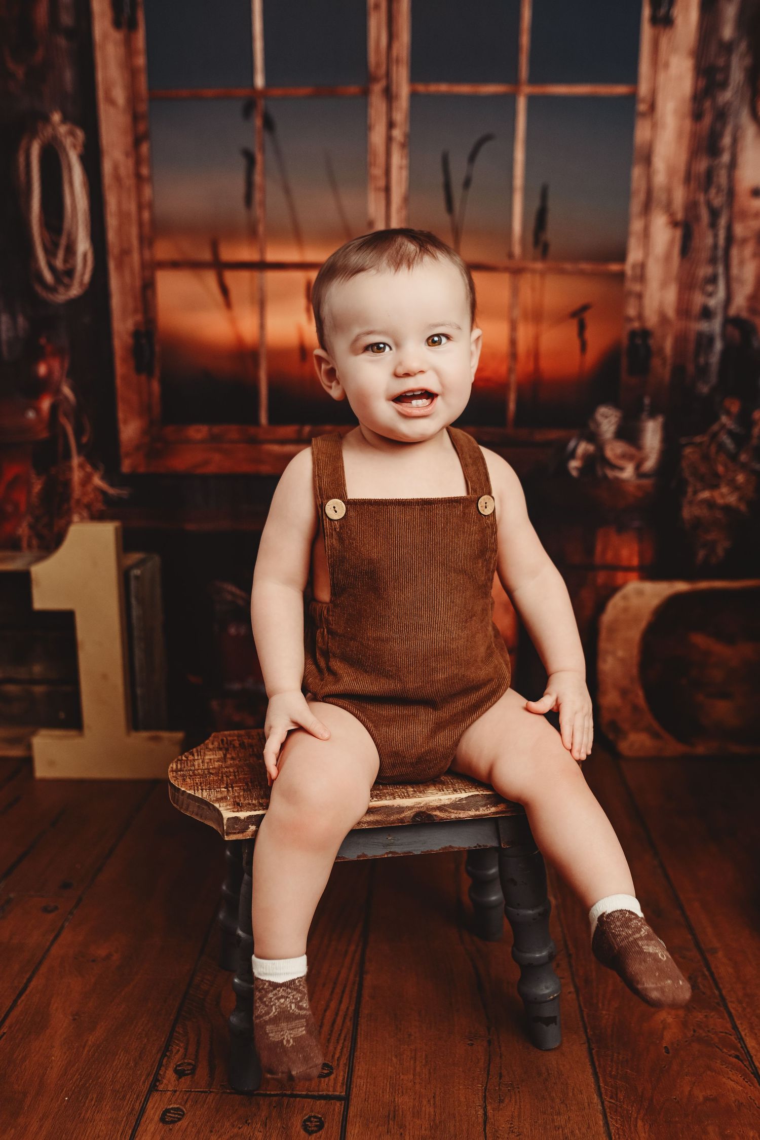 Rustic baby photography with brown overalls on wooden floor at sunset.