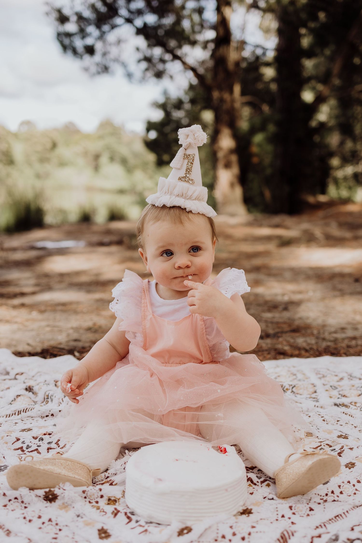 Baby in pink tulle dress and party hat sitting on blanket for first birthday photoshoot.