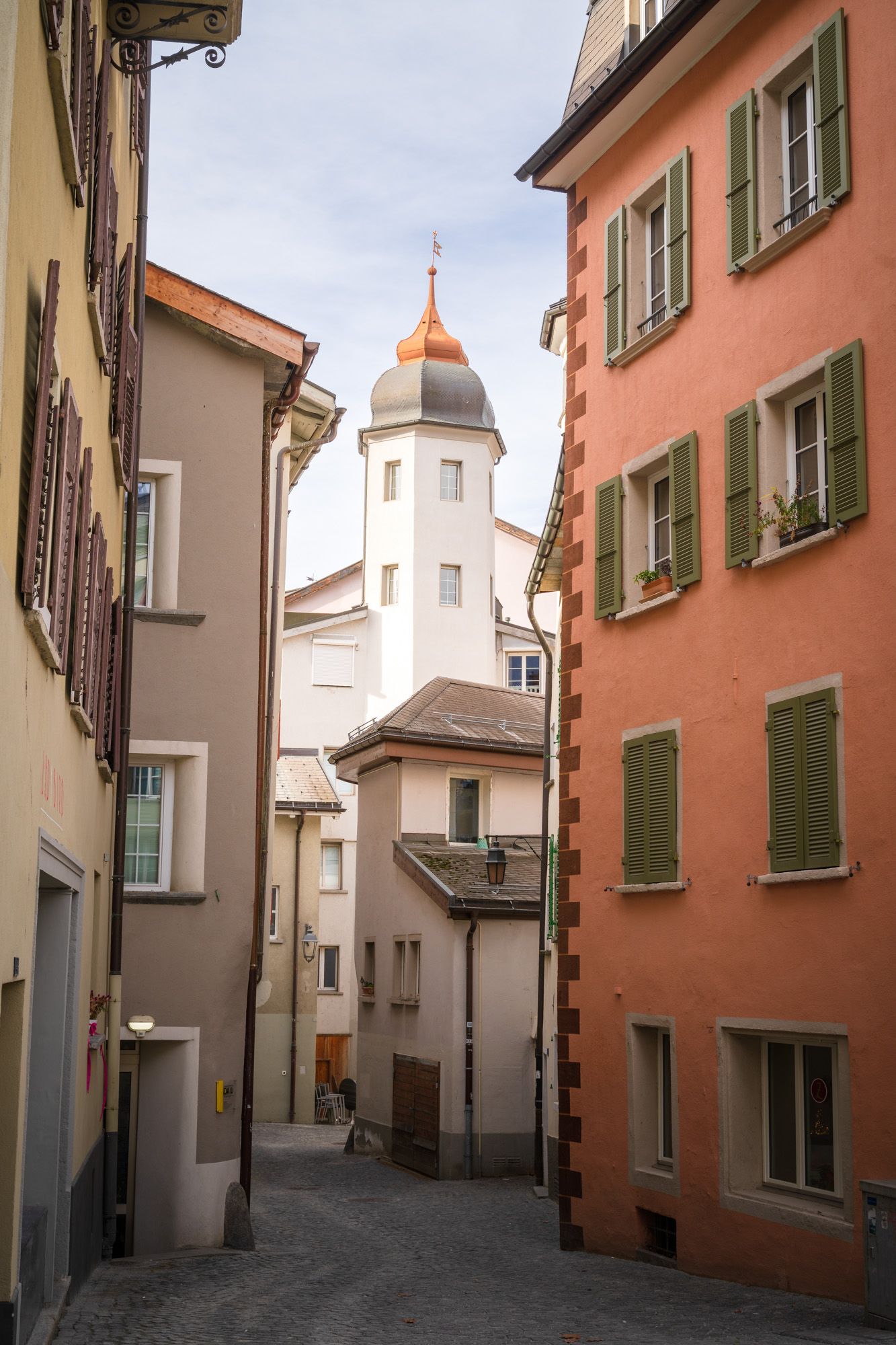 Narrow cobblestone street between colorful buildings leads to a church tower in a European town.