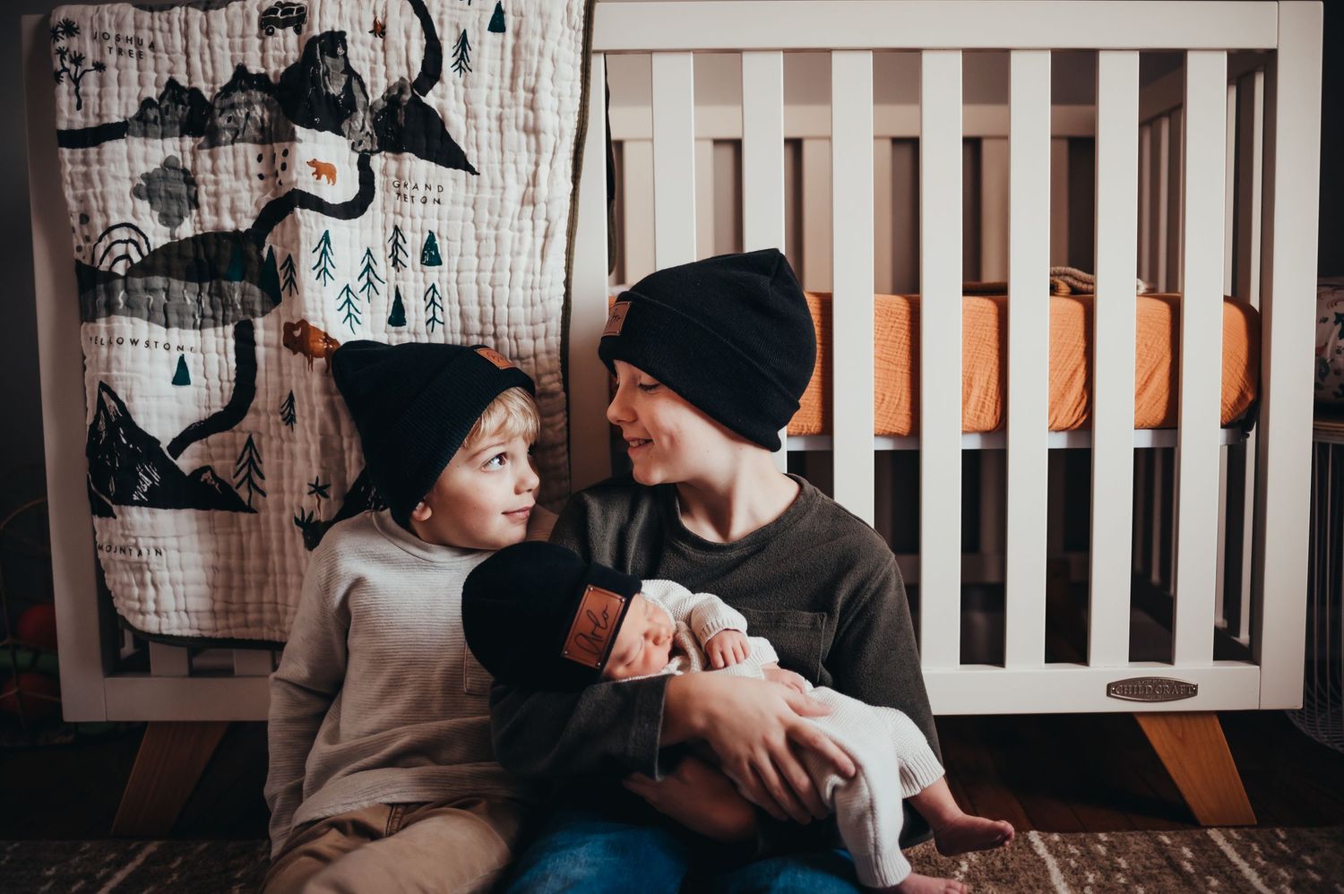 2 young boys sit on the floor in front of a crib looking at each other in matching hats and holding newborn baby brother